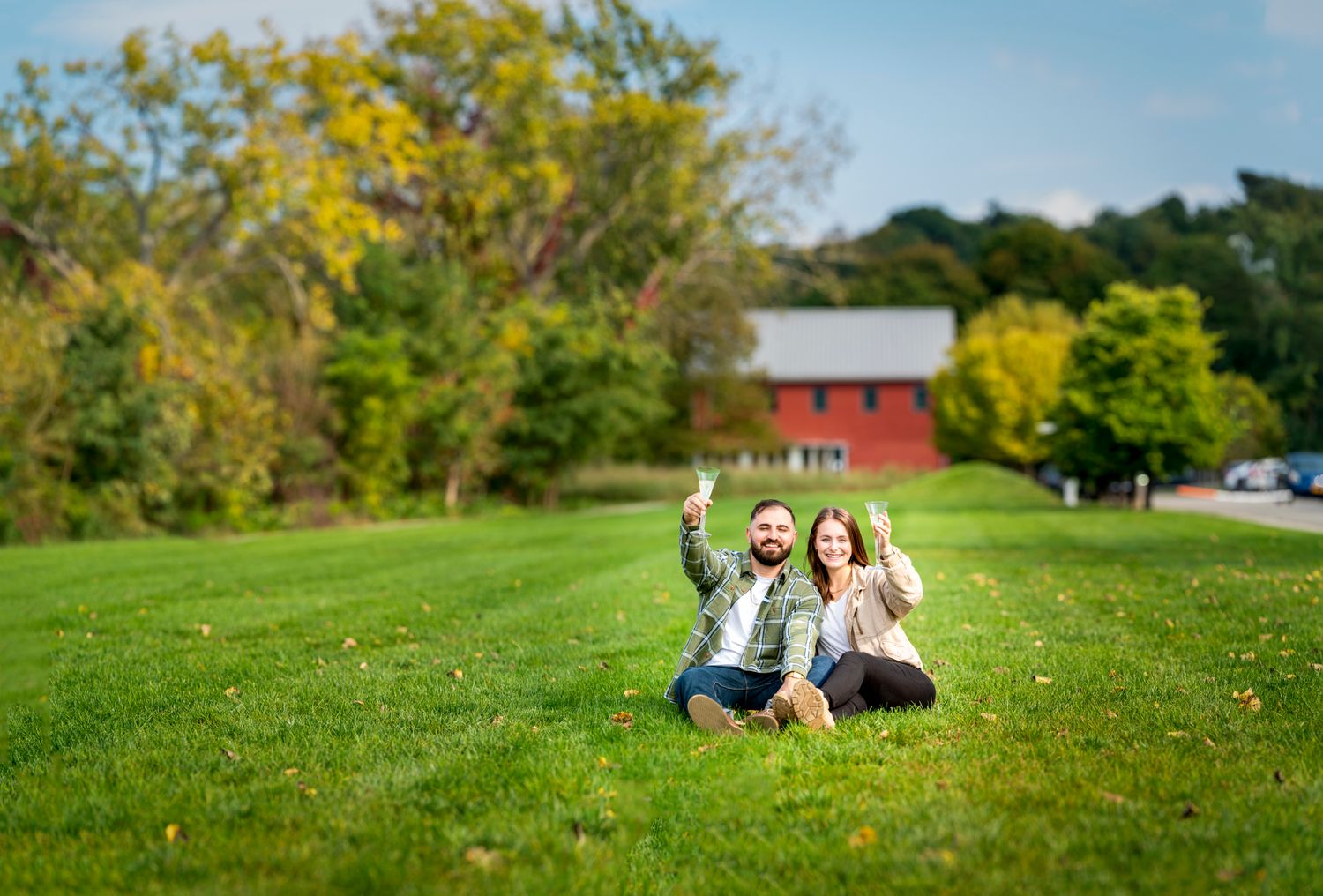 Happy couple sitting on lush green lawn giving thumbs up with red barn and trees in background.