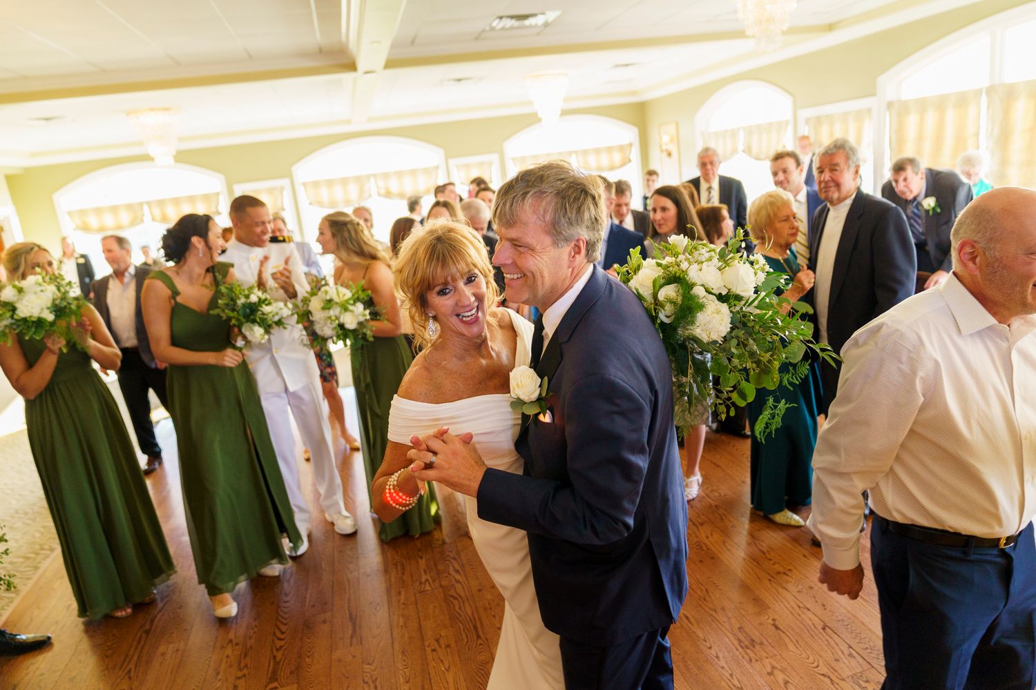 Bride and groom share a romantic first dance surrounded by guests and bridesmaids in green dresses.