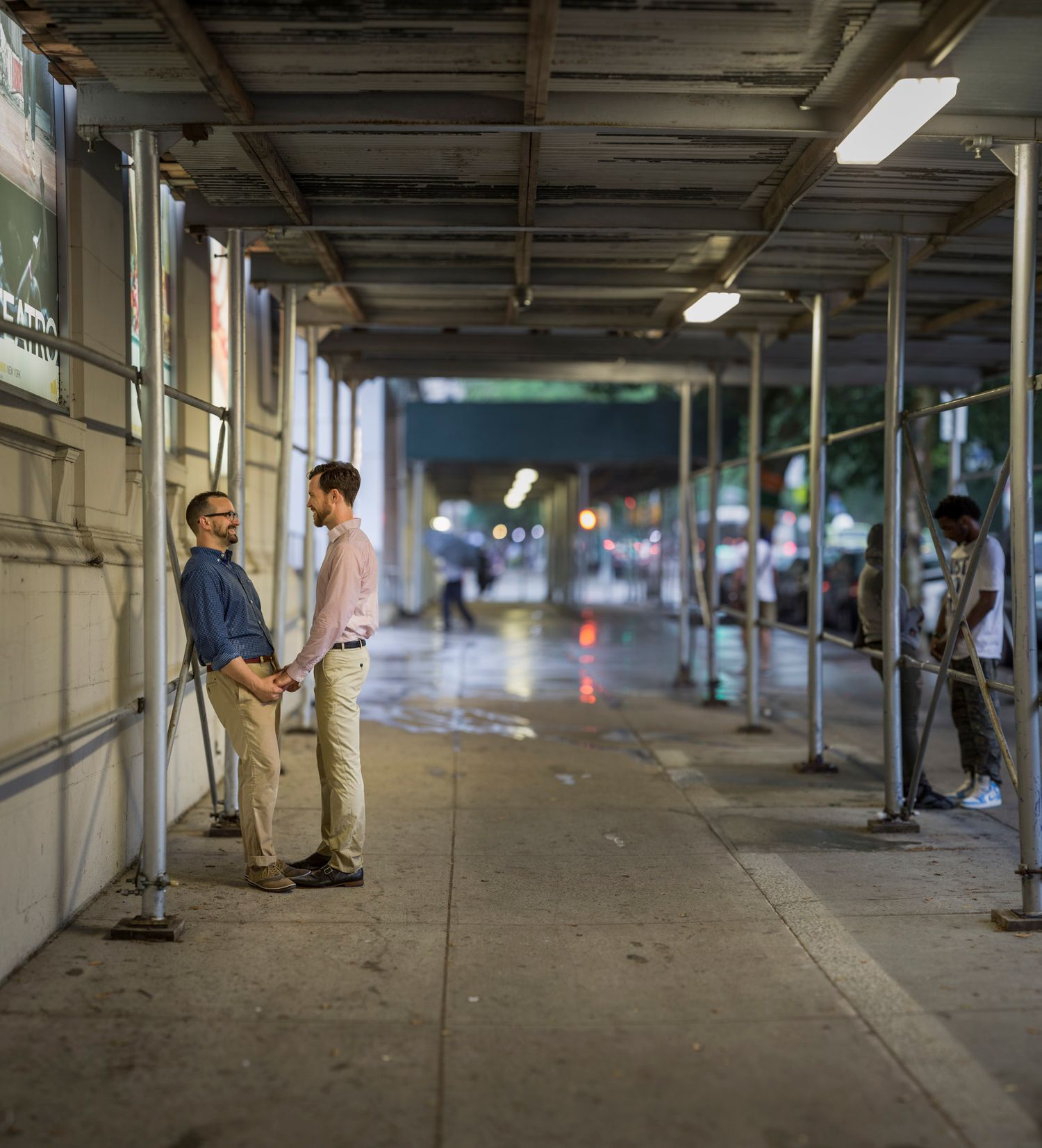 Two men stand under a covered walkway at night, with wet pavement and city lights visible in the background.