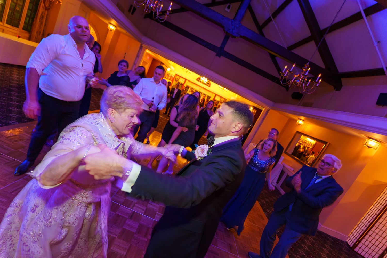 Groom dancing with elderly woman at wedding reception, guests watching in warmly lit venue.
