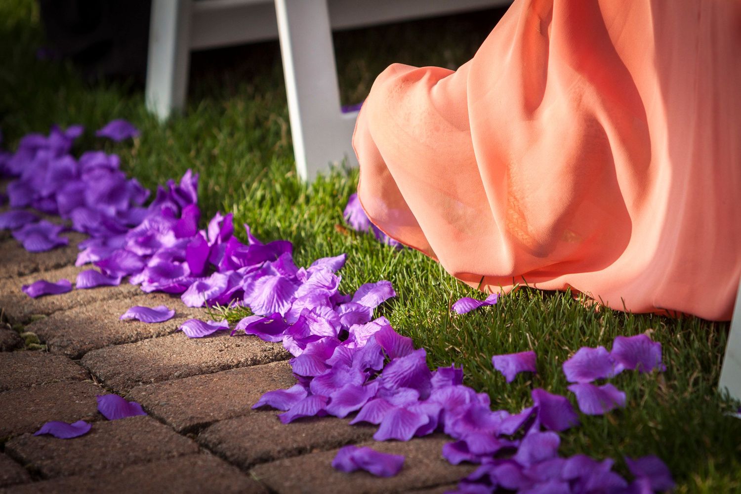 Purple flower petals scattered on brick pathway beside a flowing peach bridesmaid dress on green grass.