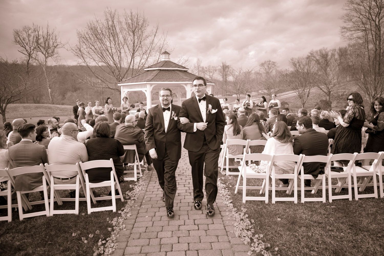 Two grooms walk down the aisle at an outdoor wedding ceremony with white chairs and a gazebo backdrop.