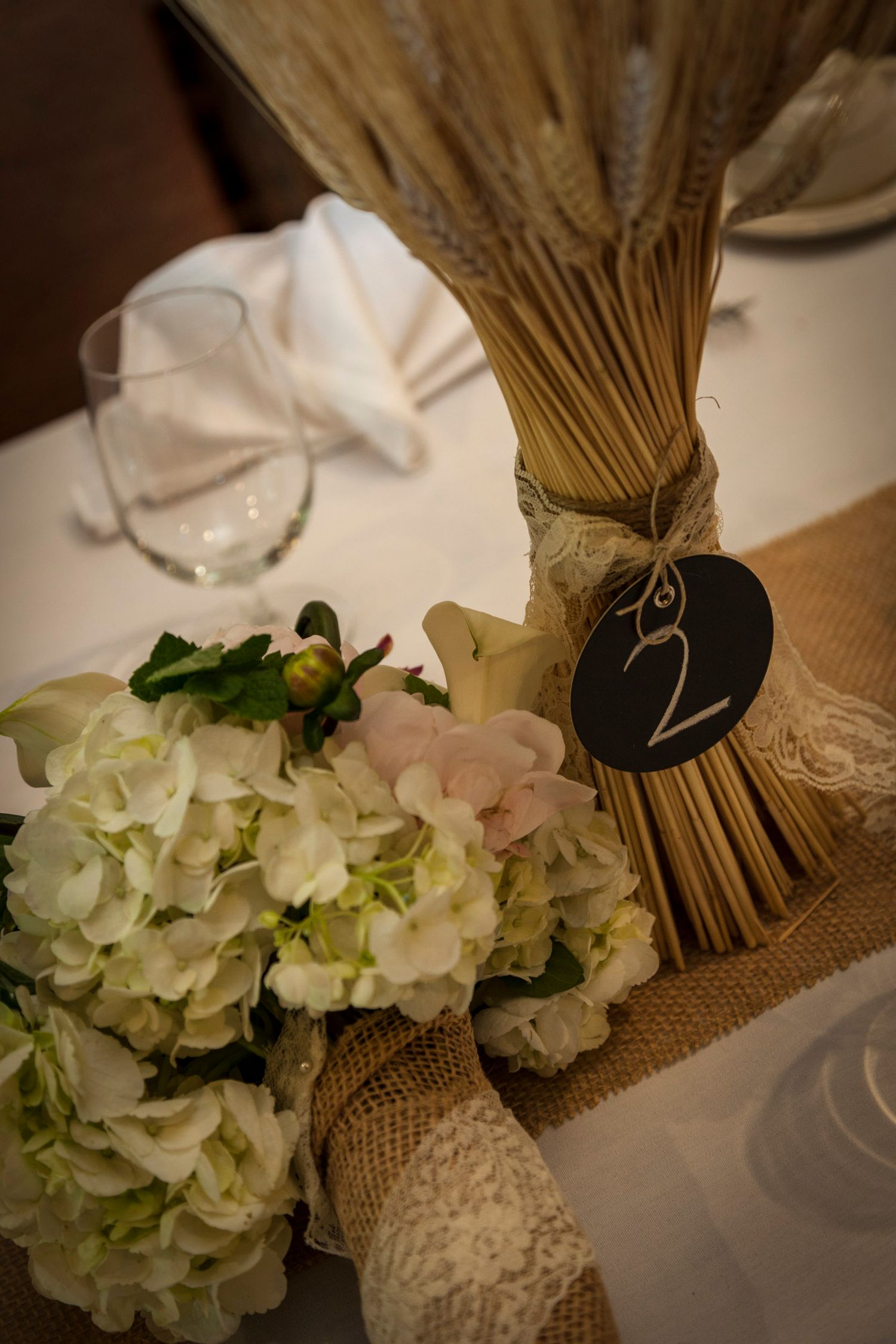 Rustic wedding table centerpiece with white hydrangeas, wheat bundle, burlap, lace, and chalkboard table number 2.