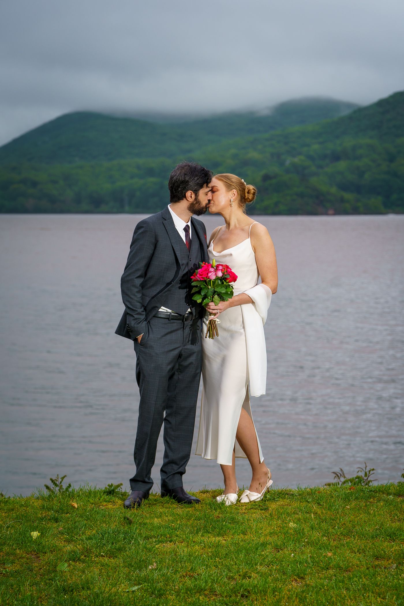 Newlywed couple kissing by a lakeside, bride in white dress holding pink roses, groom in dark suit, green hills behind.
