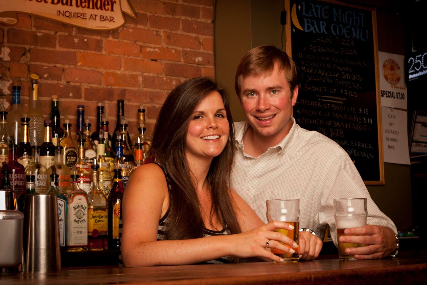 Smiling couple enjoying drinks together at a cozy bar with bottles on shelves behind them.