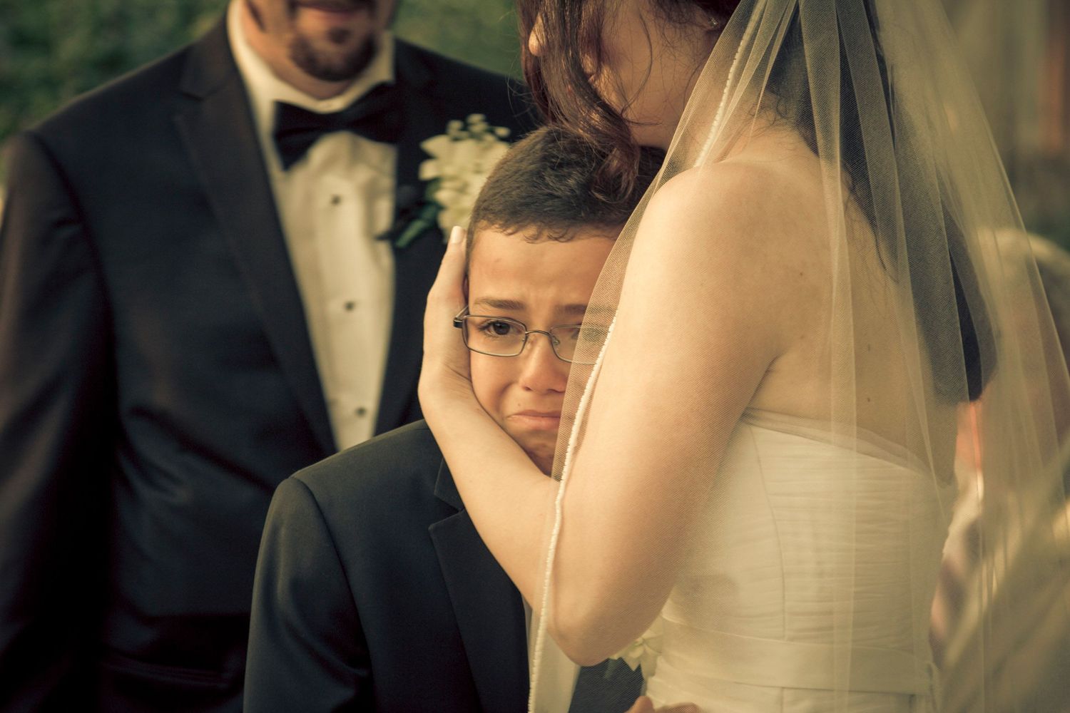 Emotional young boy hugs bride in wedding dress during outdoor ceremony.