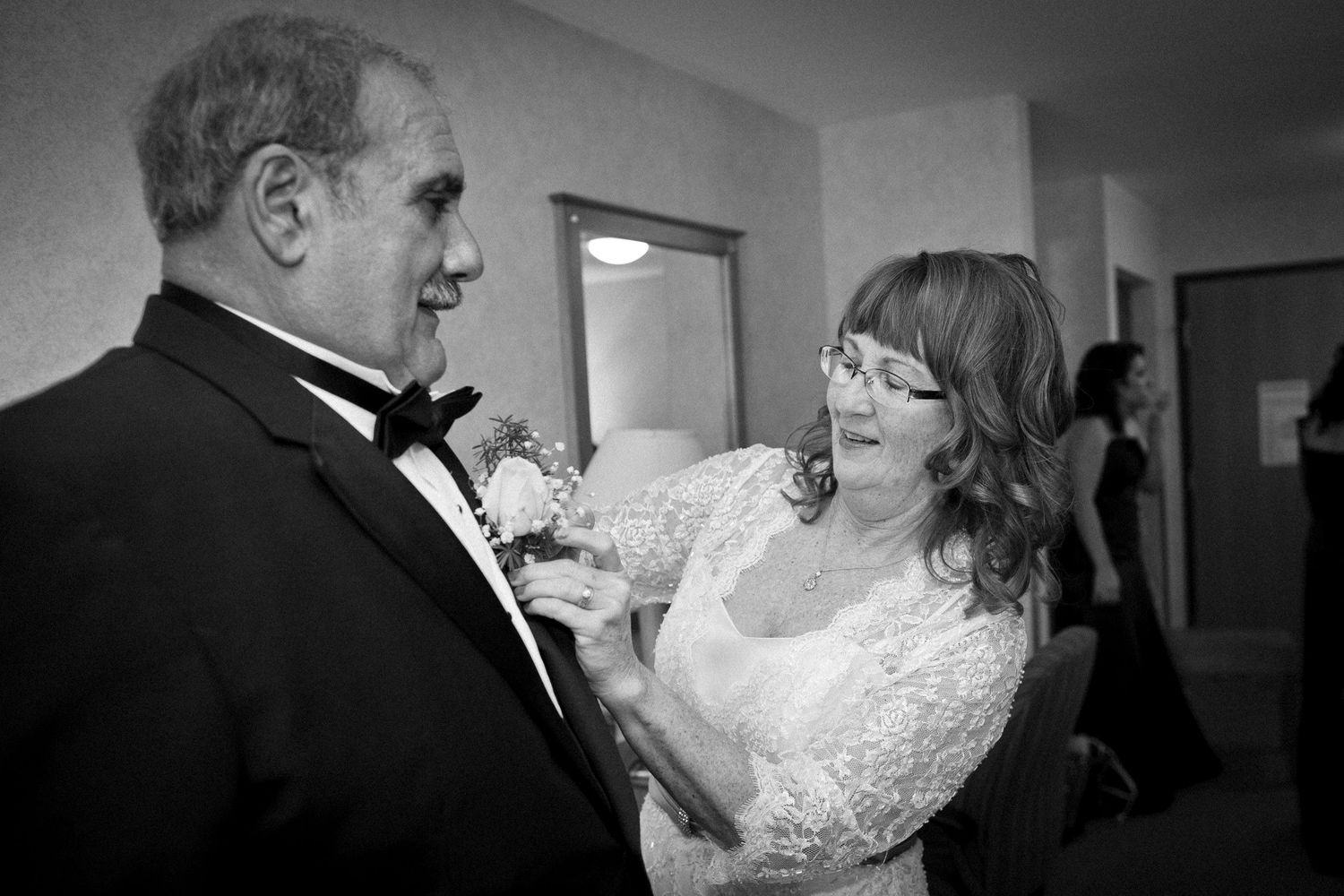 Black and white photo of a woman pinning a boutonniere on a man in a tuxedo.