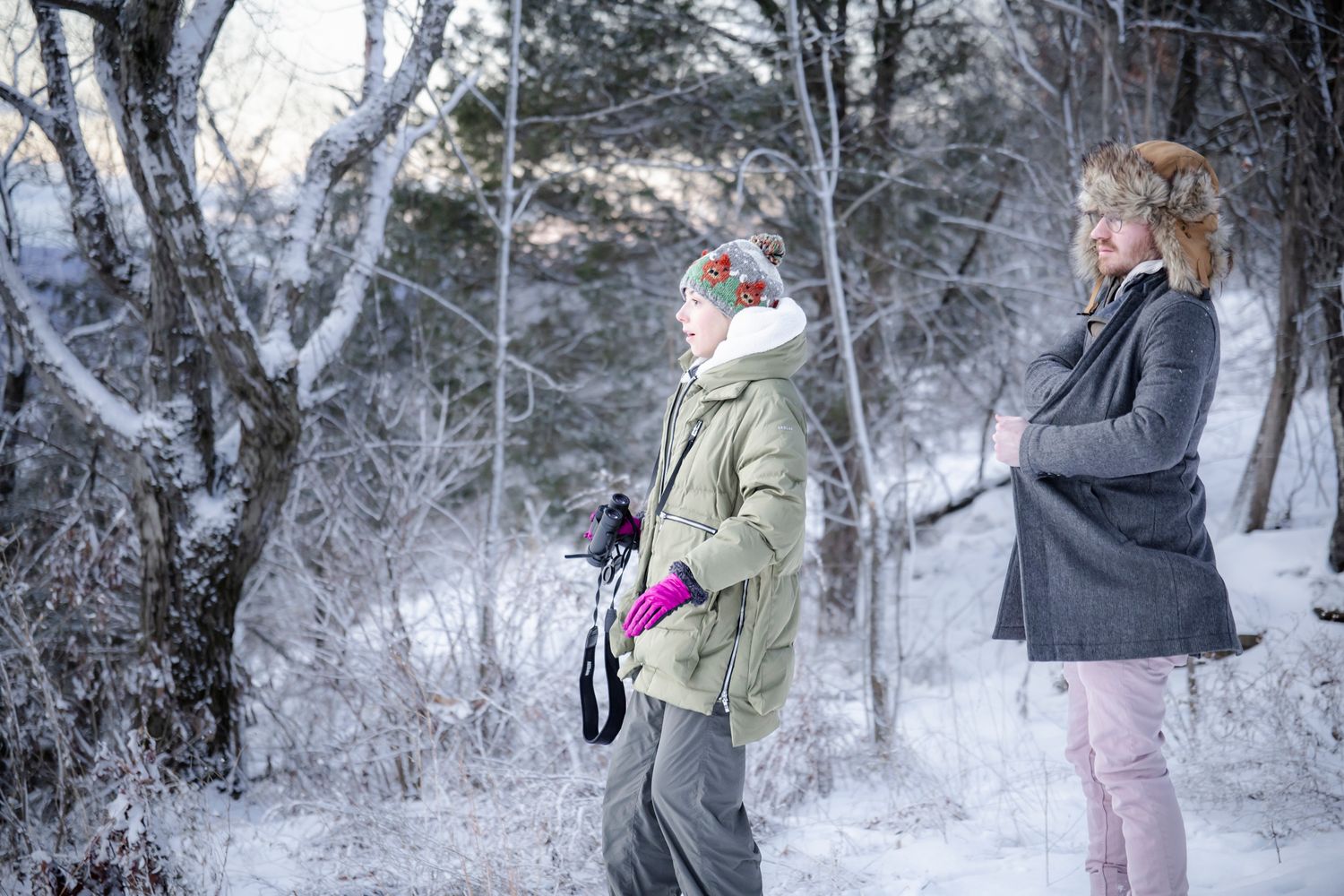 Two women stand in a snow-covered winter forest, dressed in warm coats and hats, with bare frost-covered trees behind them.