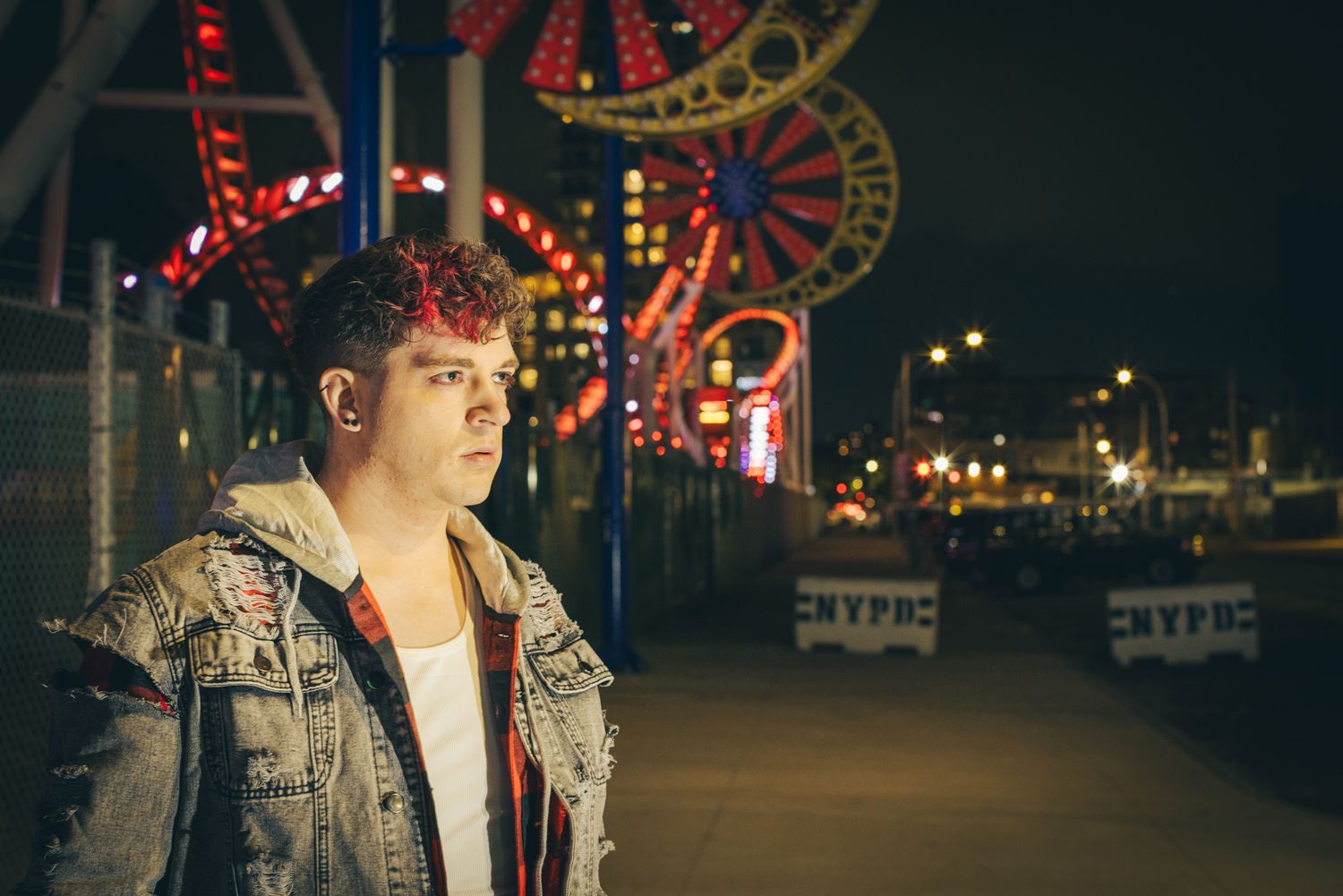 Young man with red-tinted hair in a studded denim jacket stands on a boardwalk at night, with glowing amusement park rides behind him.
