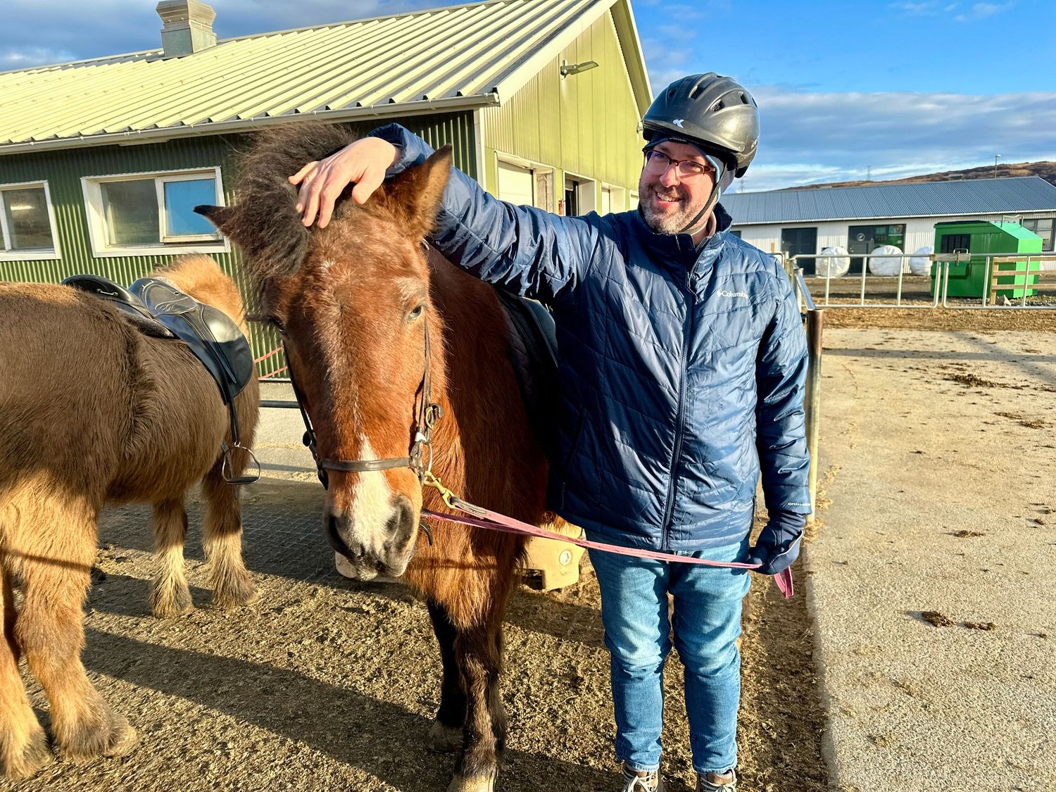 Smiling man in riding helmet petting brown horse at equestrian stable on sunny day.