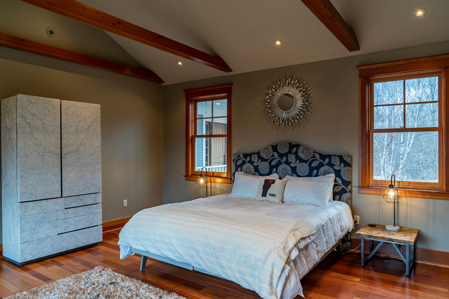 Cozy bedroom with vaulted wood beam ceiling, gray walls, queen bed, hardwood floors, and large windows with warm natural light.