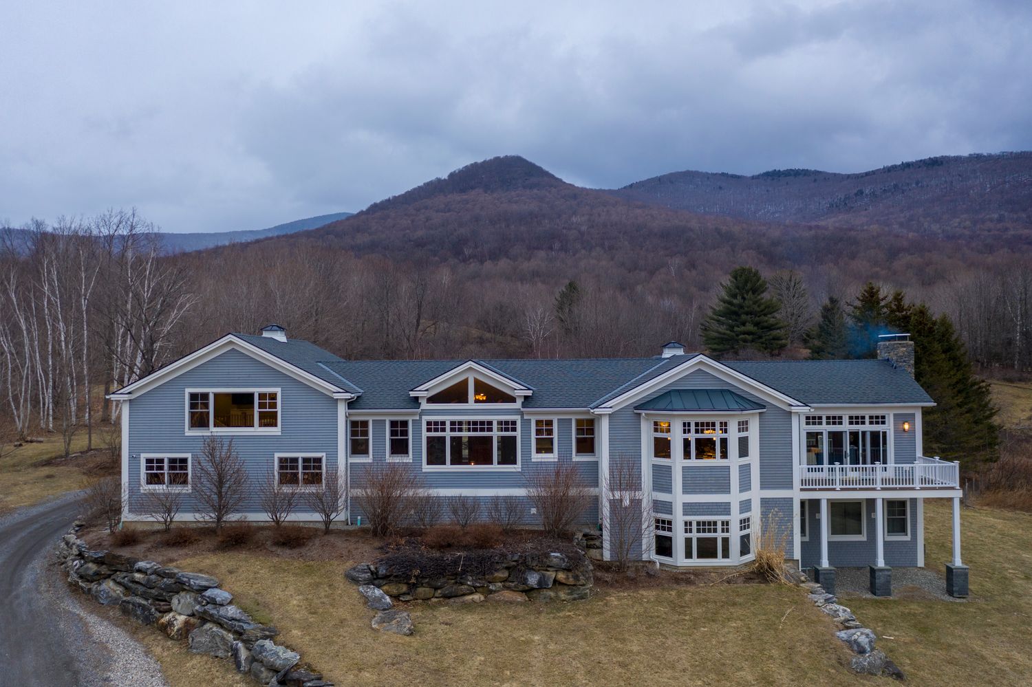 Large blue colonial home with wraparound deck nestled against a scenic mountain backdrop in a rural New England setting.