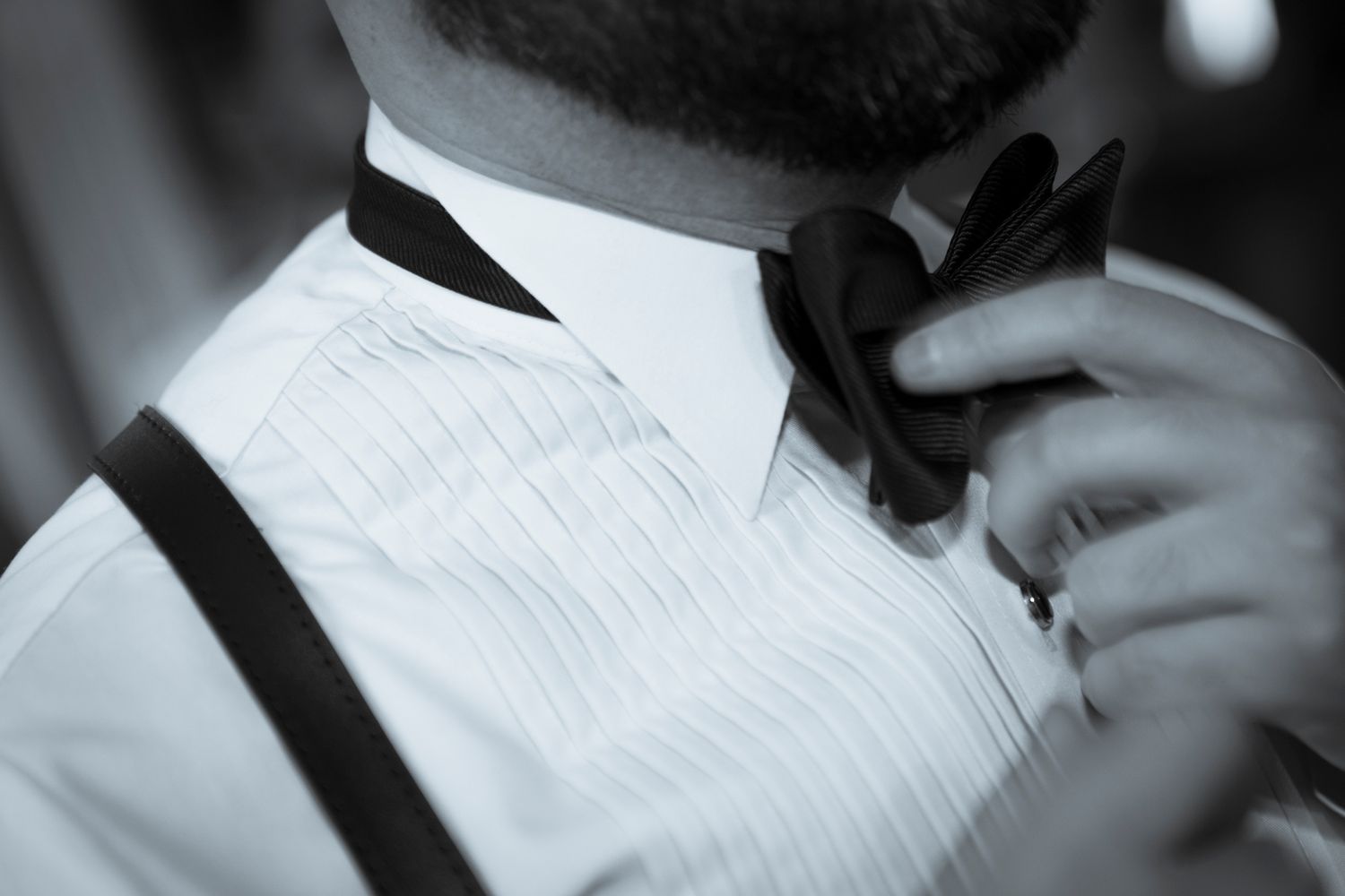 Groom adjusting black bow tie wearing white tuxedo shirt and suspenders in black and white photo.