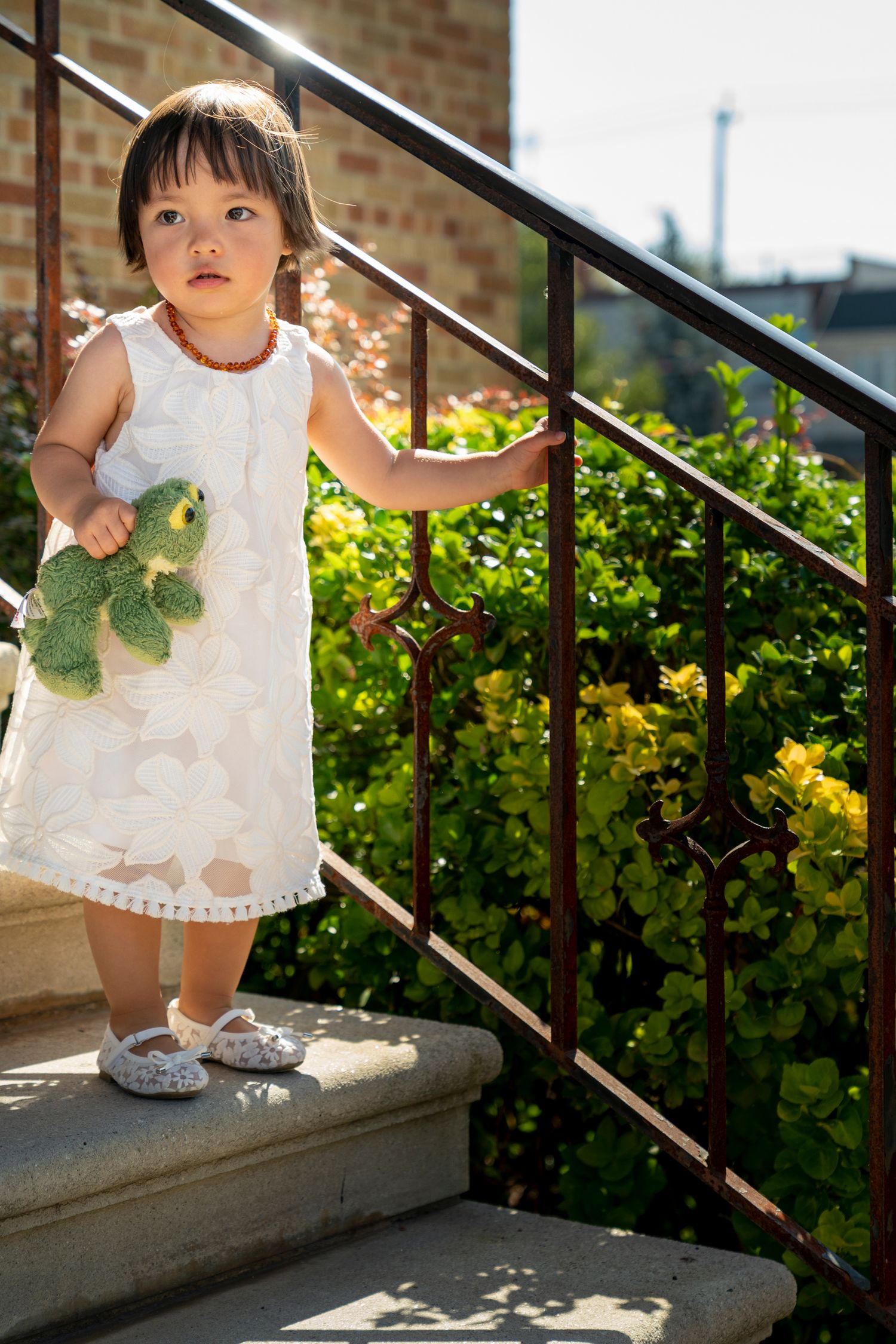 Young girl in white dress holding stuffed turtle toy on sunlit outdoor staircase with green foliage.