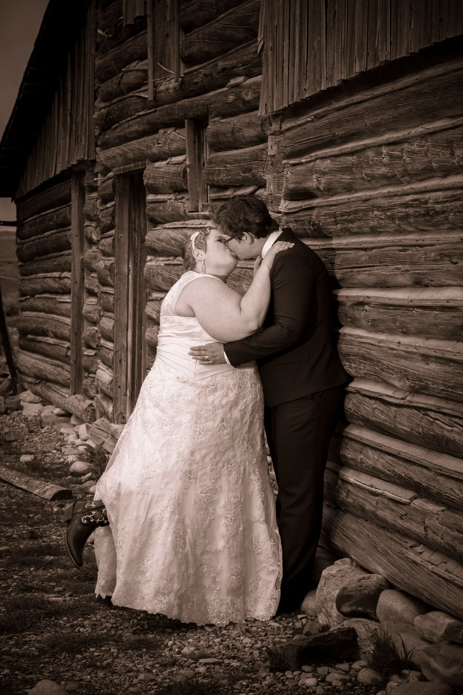 Bride and groom share a romantic kiss in sepia tone beside a rustic log cabin wall on their wedding day.