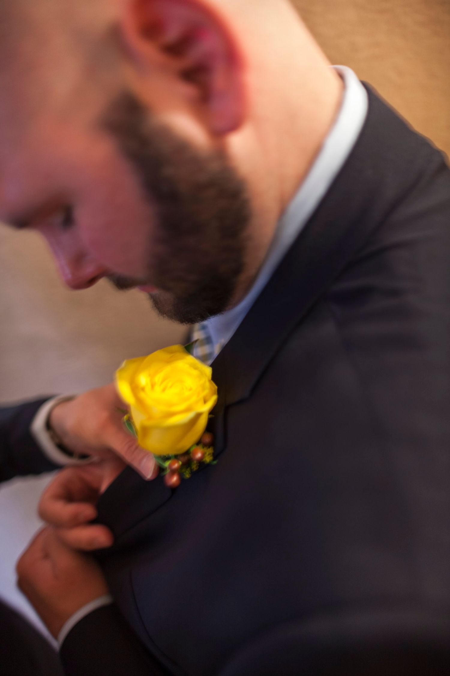 A bearded groom in a dark suit pins a yellow rose boutonniere to his lapel at a wedding ceremony.