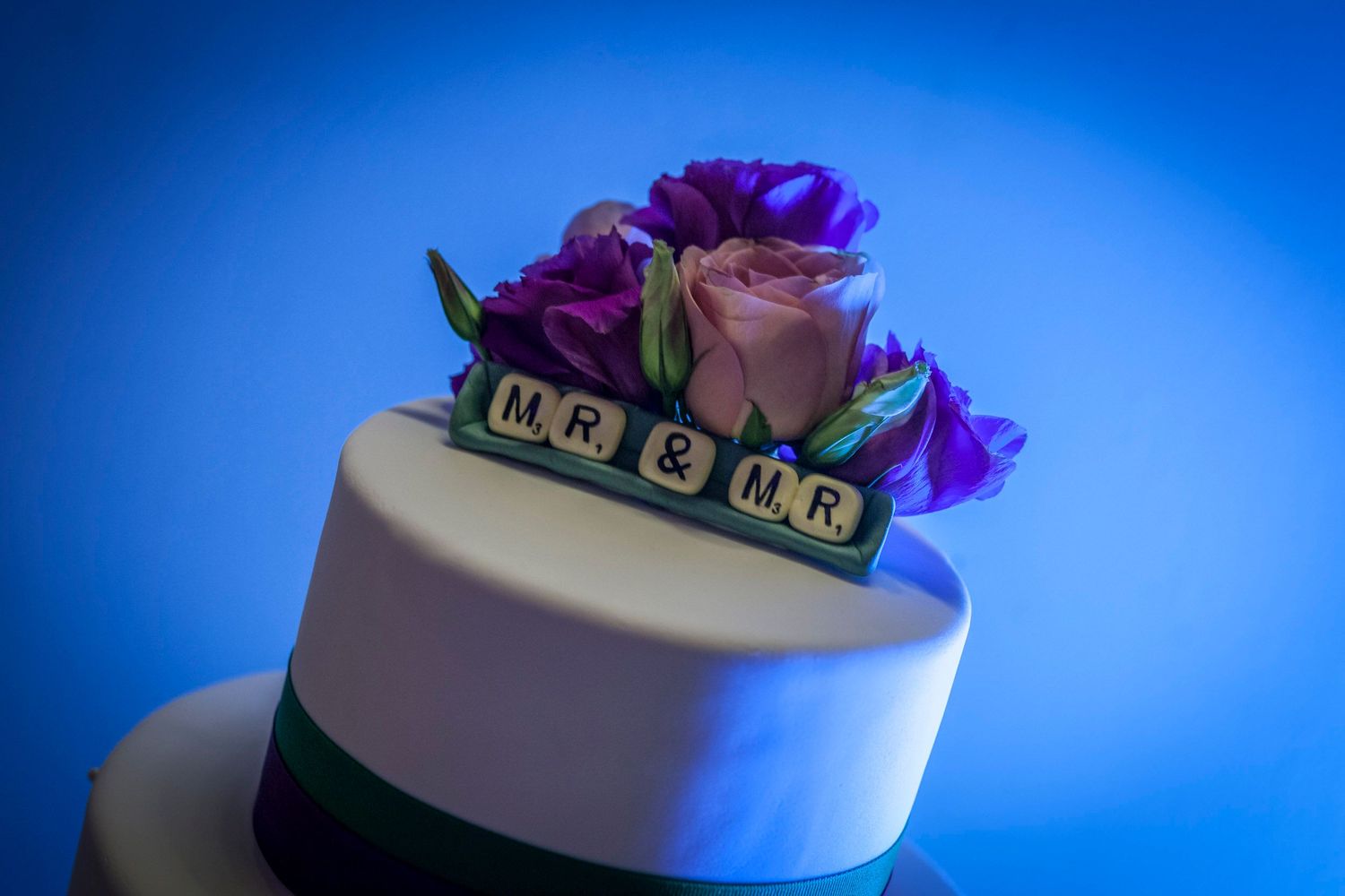 Wedding cake topper with Scrabble tiles spelling Mr & Mr adorned with purple roses on a blue background.