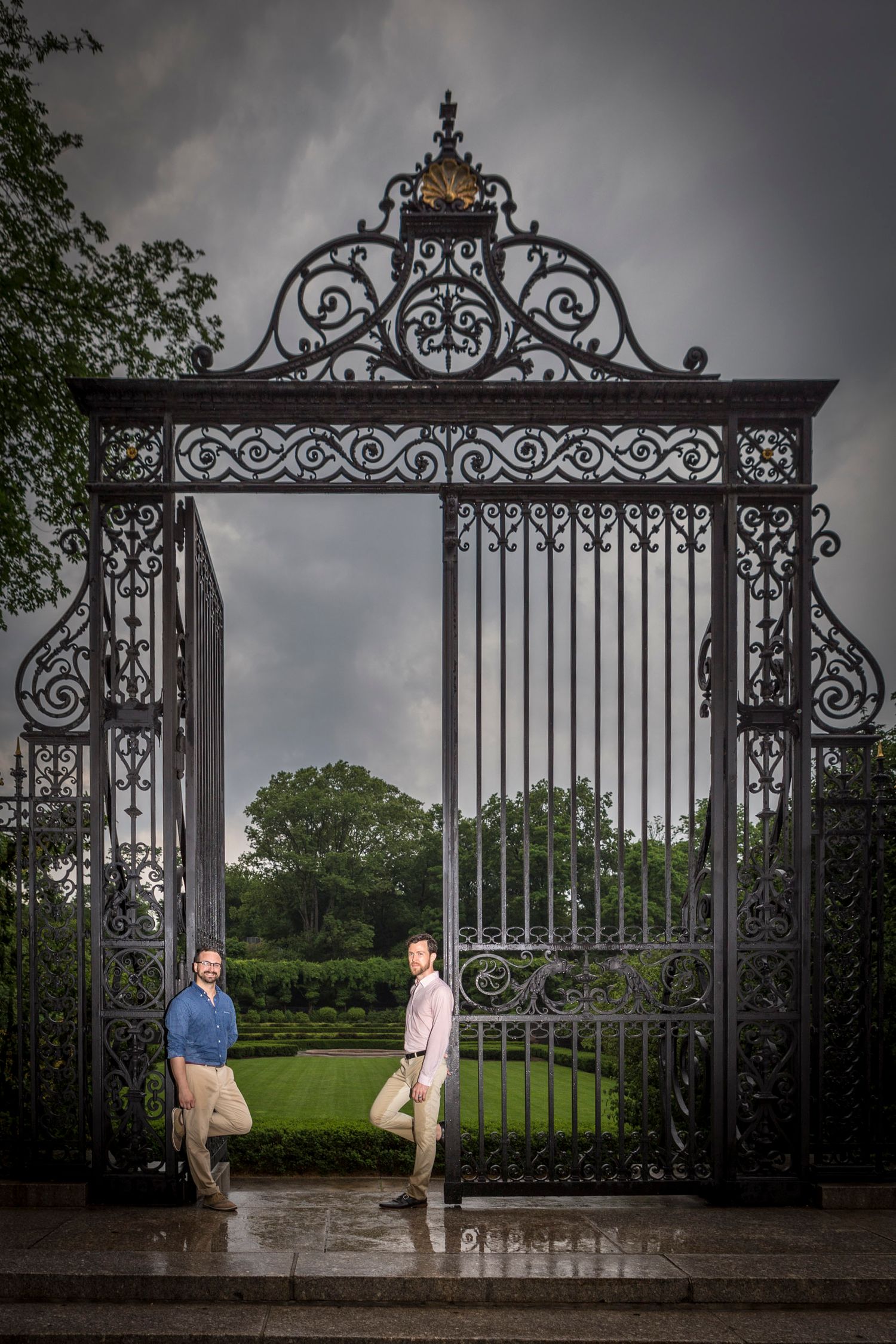 Two men posing at an ornate wrought iron gate in a lush garden under dramatic stormy skies.