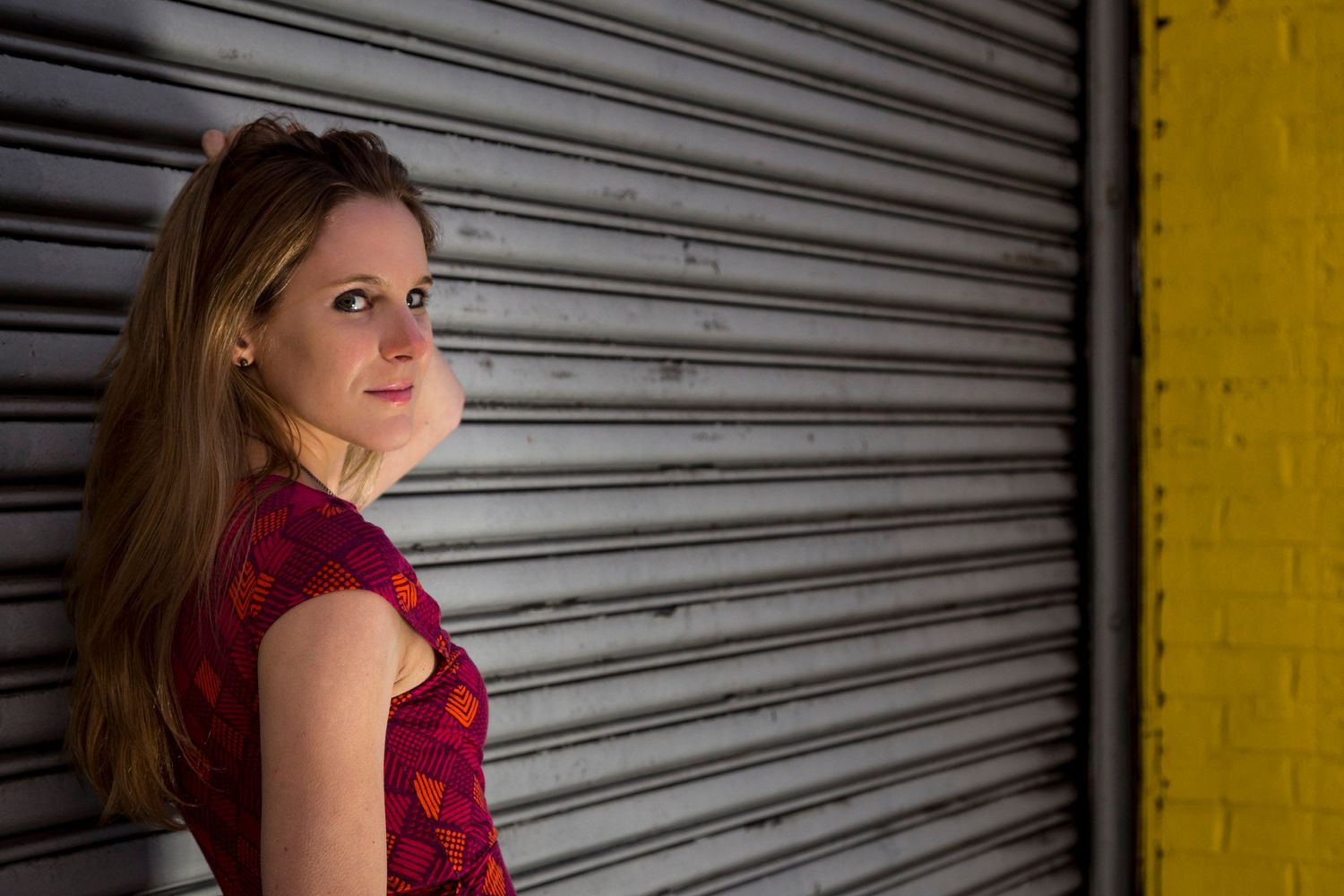 Young woman in red floral dress posing against a metal roller shutter door next to a yellow wall.
