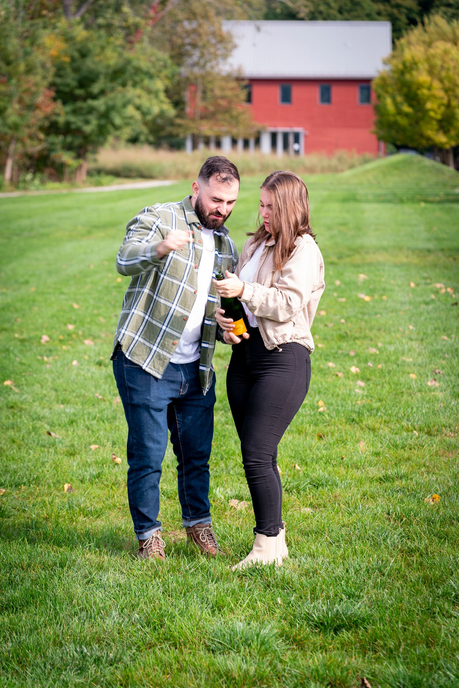 Couple opening a champagne bottle outdoors on green grass with a red barn in the background.