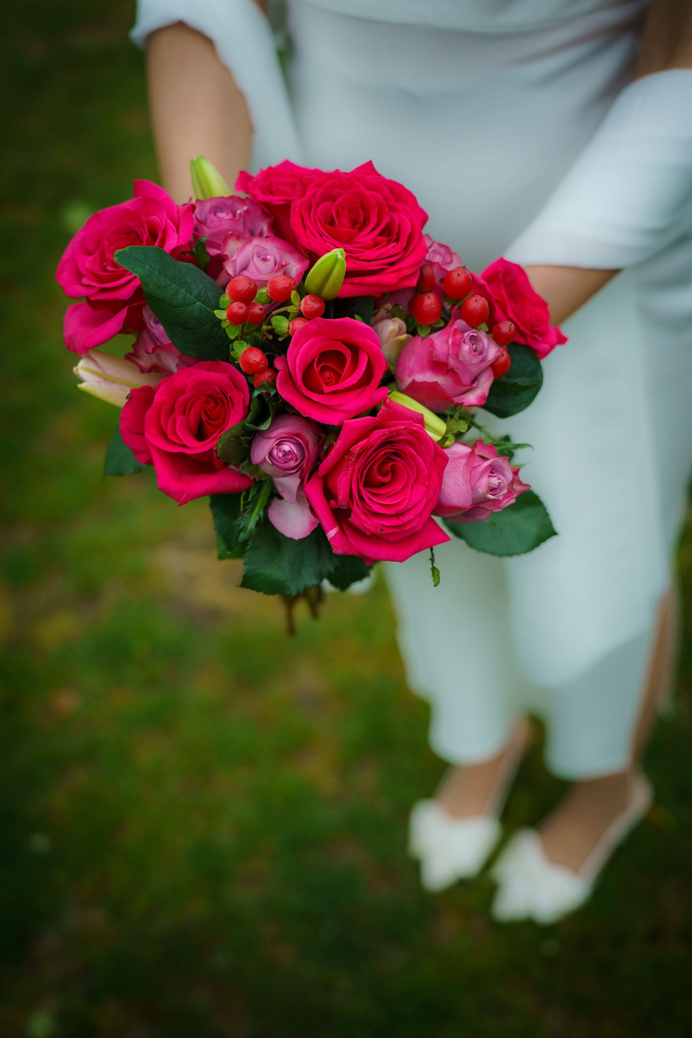 Bride in white dress holding a vibrant bouquet of red and pink roses with green leaves outdoors.