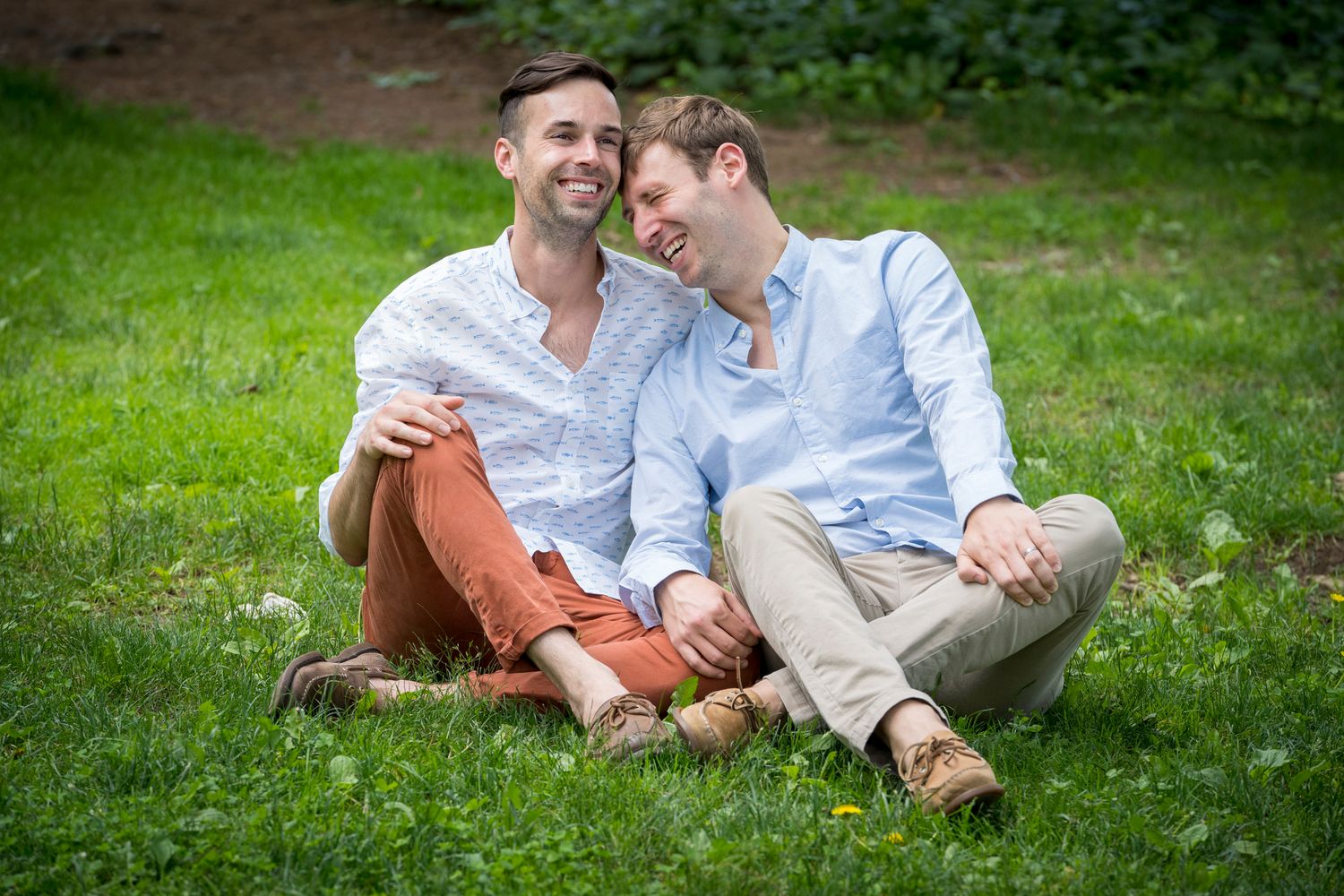 Two men sitting together on green grass, smiling and laughing during an outdoor engagement photo session.