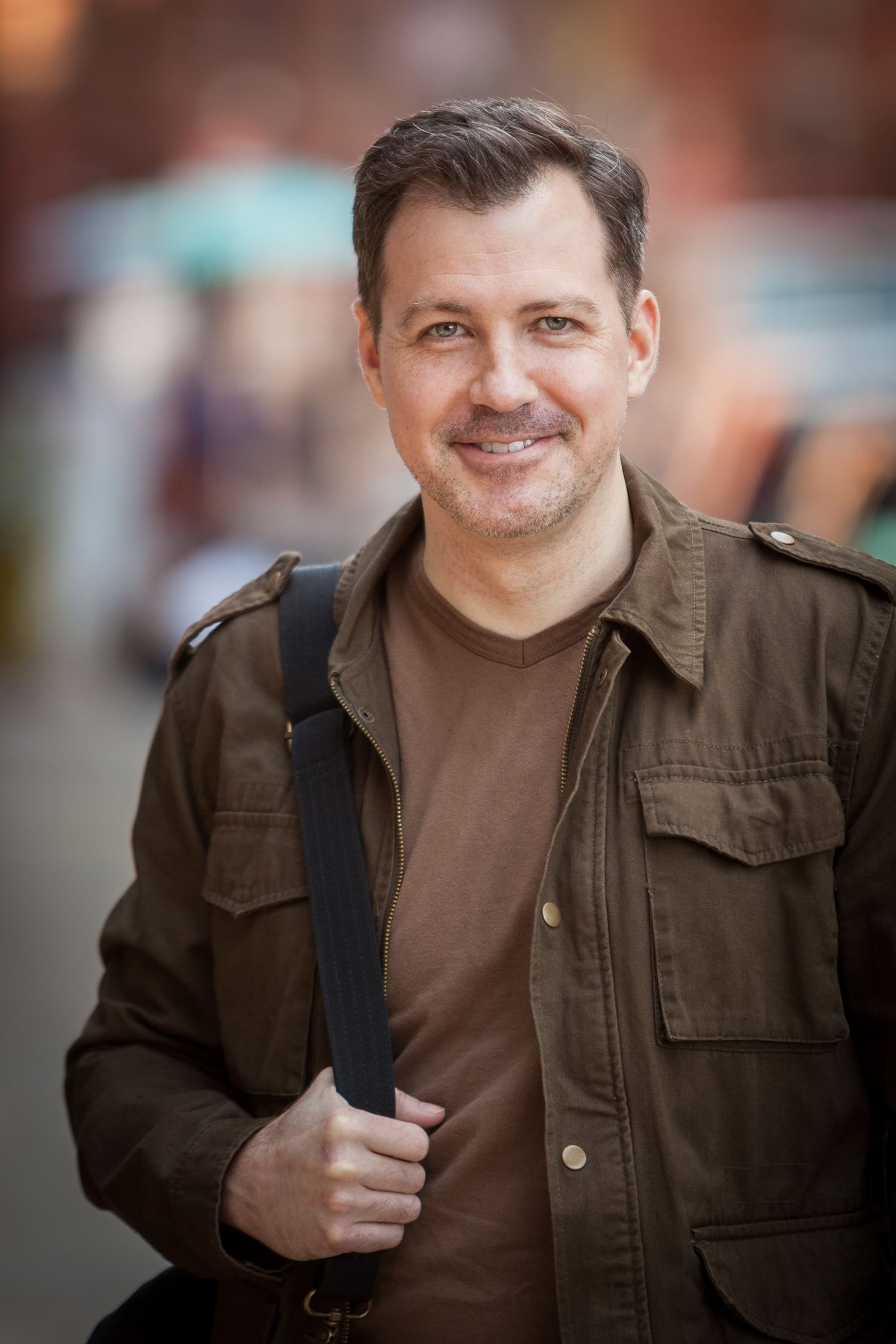 Smiling middle-aged man in brown jacket carrying bag on city street.