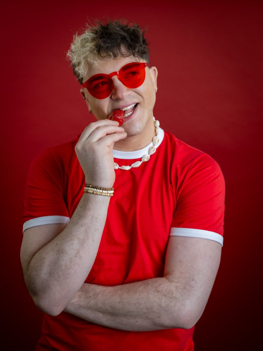 Man in red t-shirt and red heart sunglasses eating a strawberry against a red background.