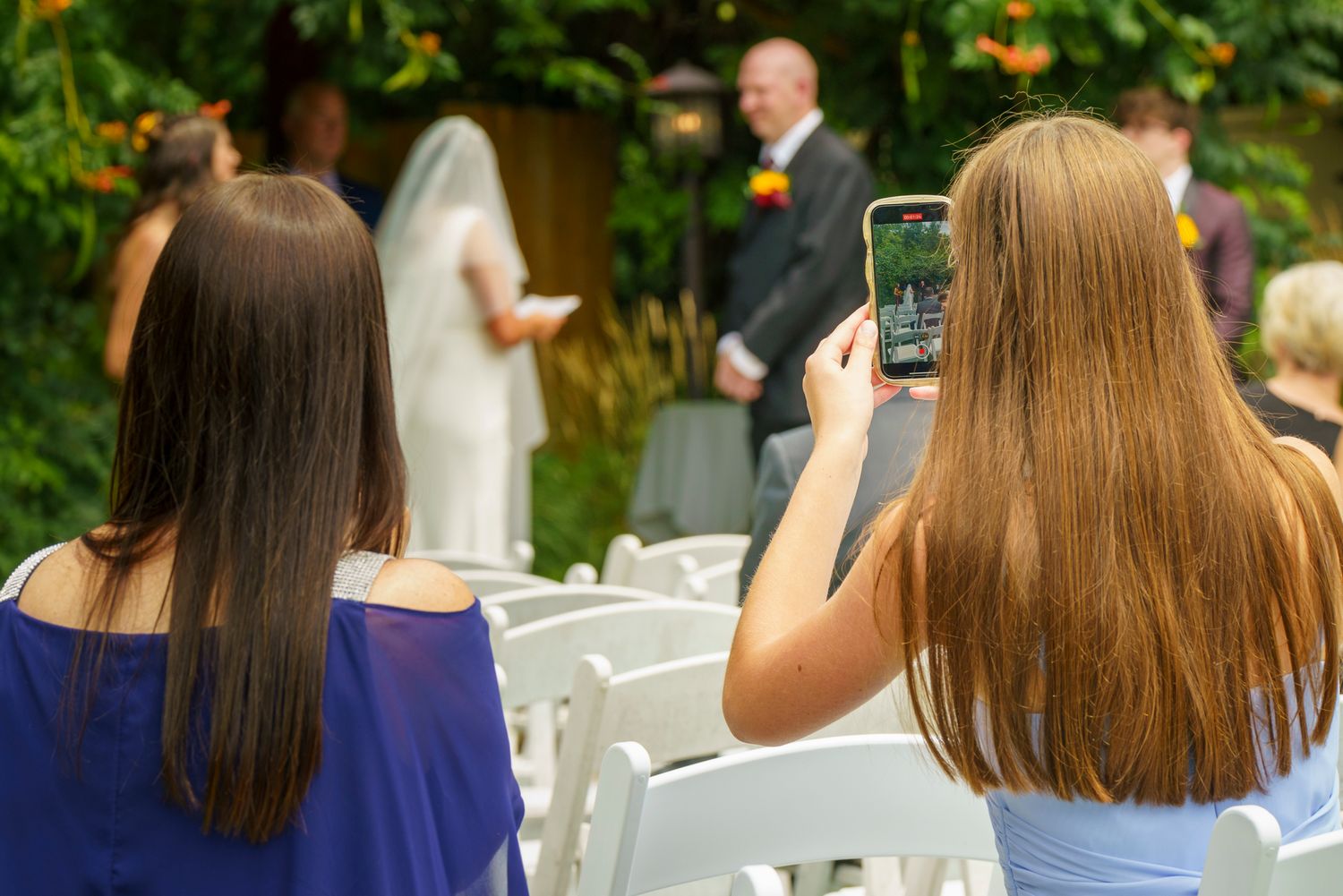 Two women with long brown hair photograph a wedding ceremony outdoors, one using a smartphone to capture the bride and groom.