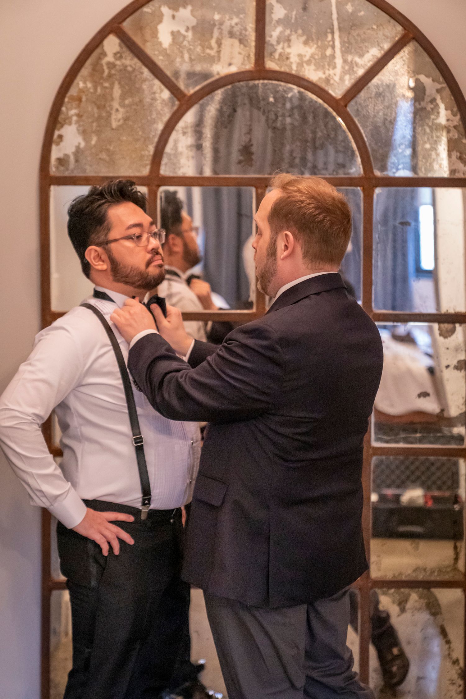 A man in a suit adjusts another man's tie near a vintage arched mirror in an elegant indoor setting.