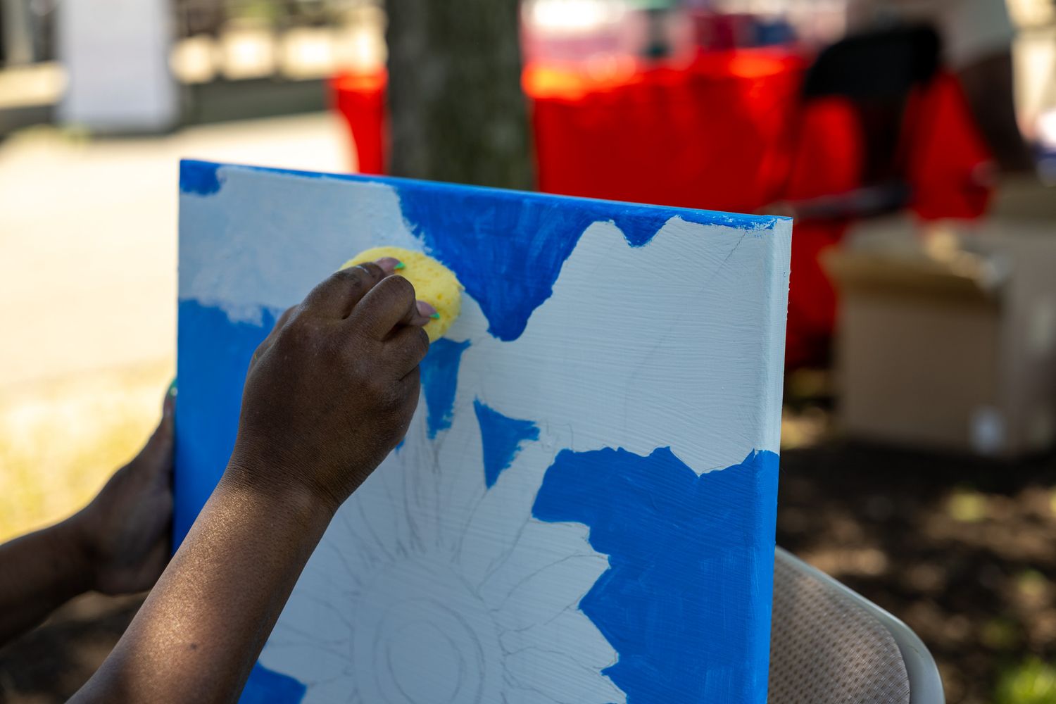Person painting blue and white floral design on canvas outdoors during a community art event.