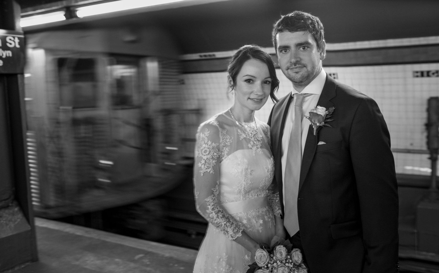 Bride and groom pose at NYC subway station in wedding attire, with blurred train passing behind them.