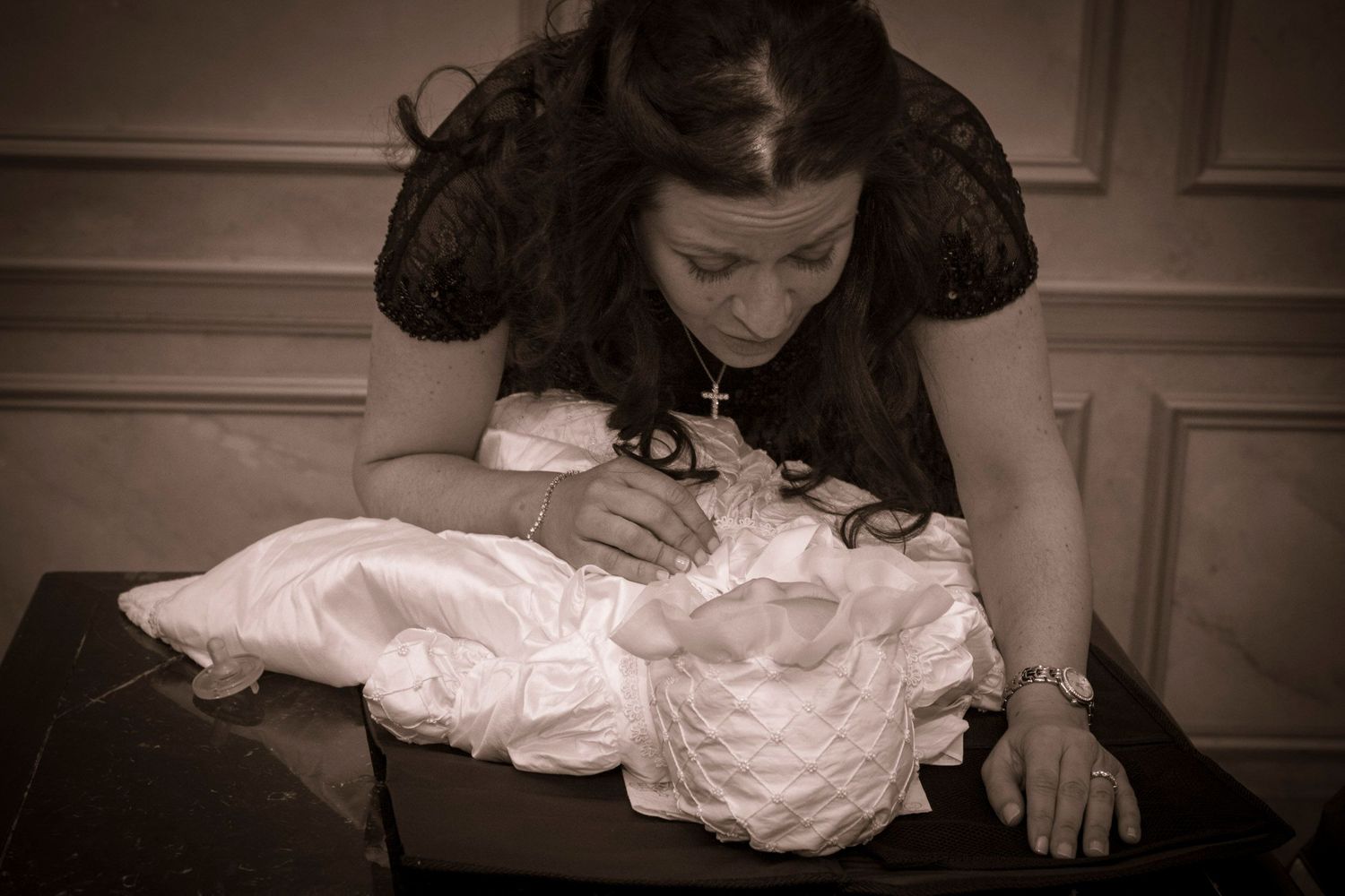 Woman in black dress leaning over white christening gown laid on dark surface, wearing cross necklace.