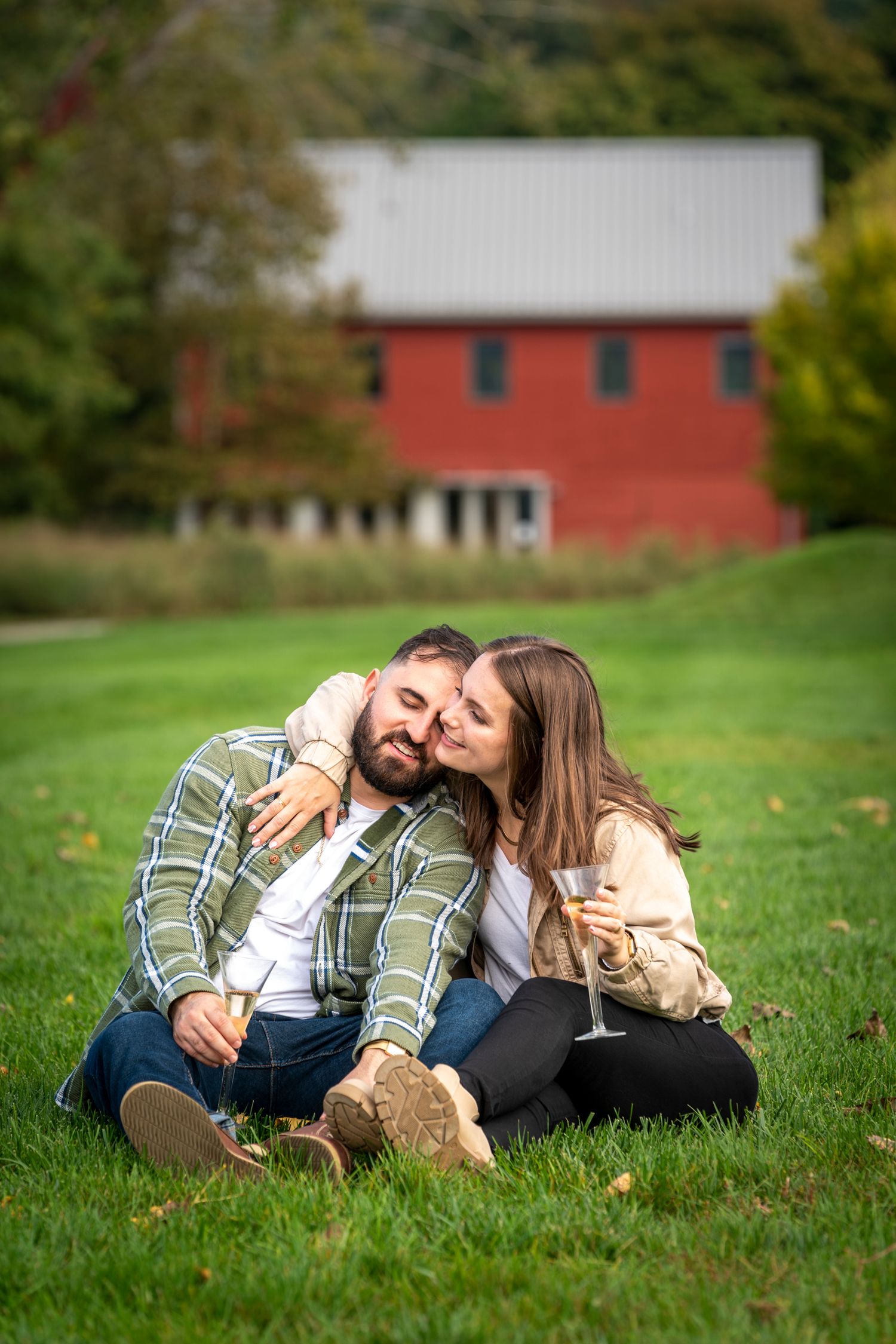 Couple sitting on green grass sharing a romantic moment near a red barn.