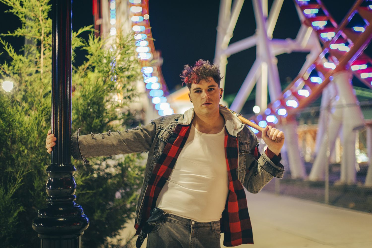Smiling young man in a plaid flannel and white tee leans on a black lamppost with a bat on his shoulder near a lit carnival ride.