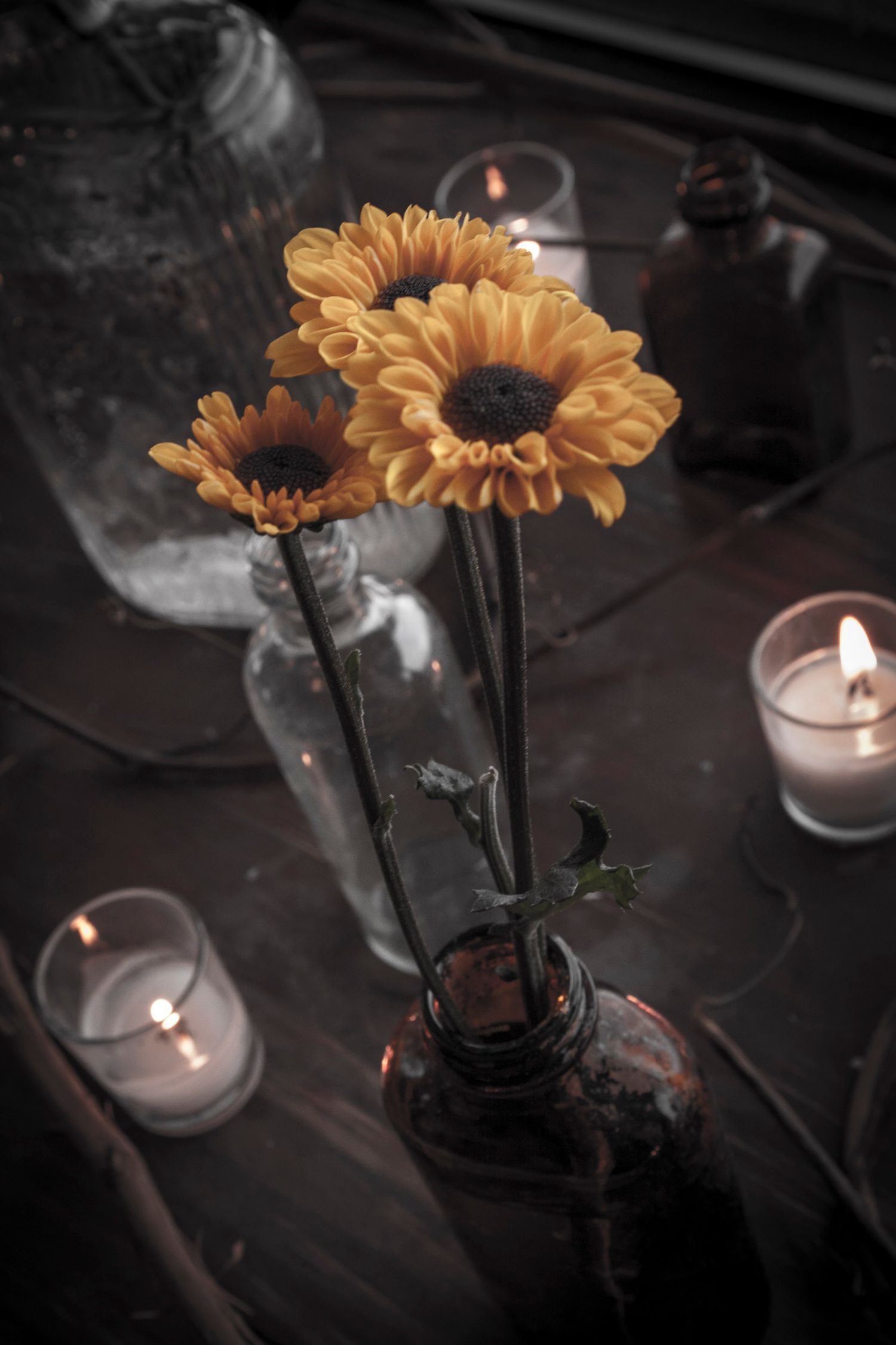 Yellow gerbera daisies in amber glass bottle vase surrounded by lit candles on dark rustic table.