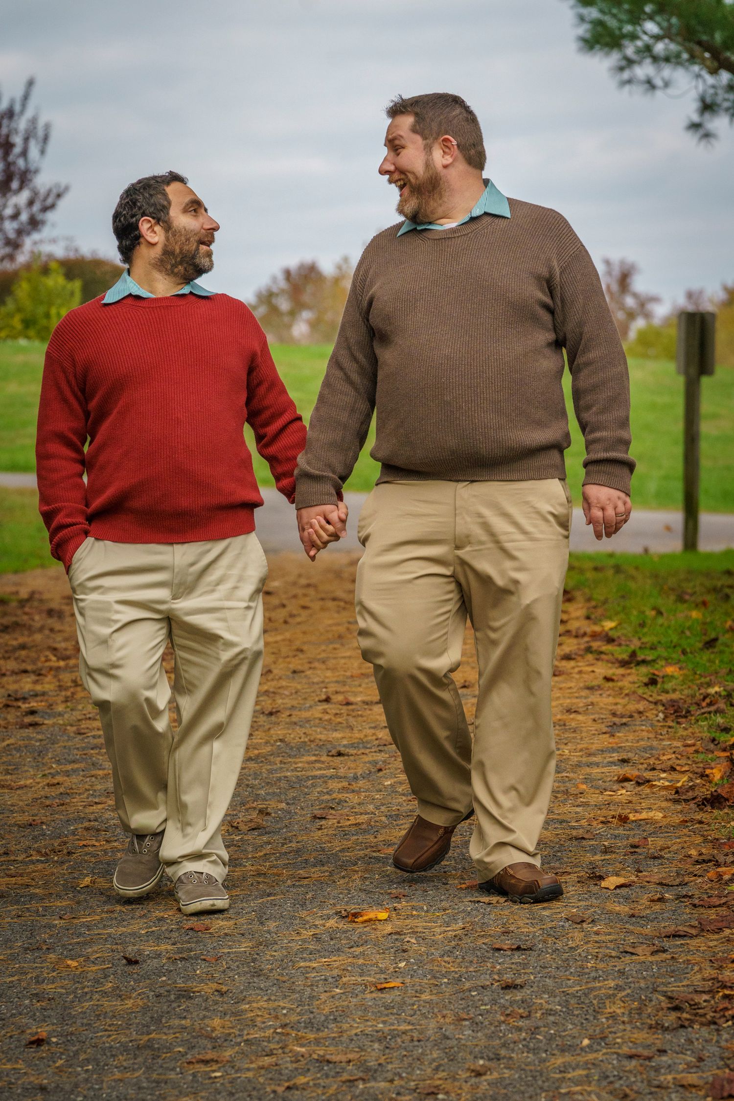 Two men holding hands and smiling while walking together on a park path in autumn, wearing sweaters and khaki pants.