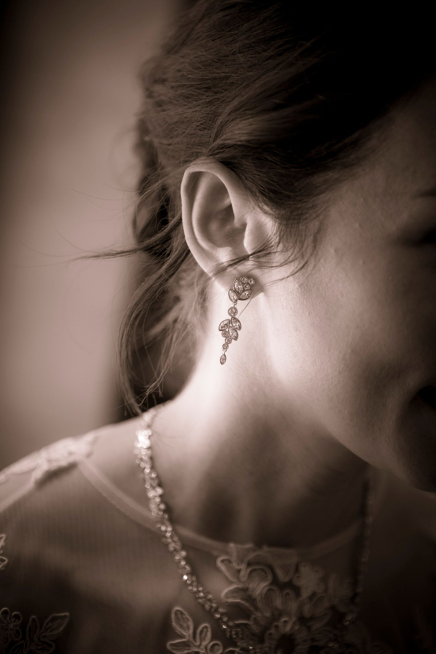 Sepia-toned close-up of a bride wearing an elegant chandelier earring and lace dress, showcasing delicate wedding jewelry.