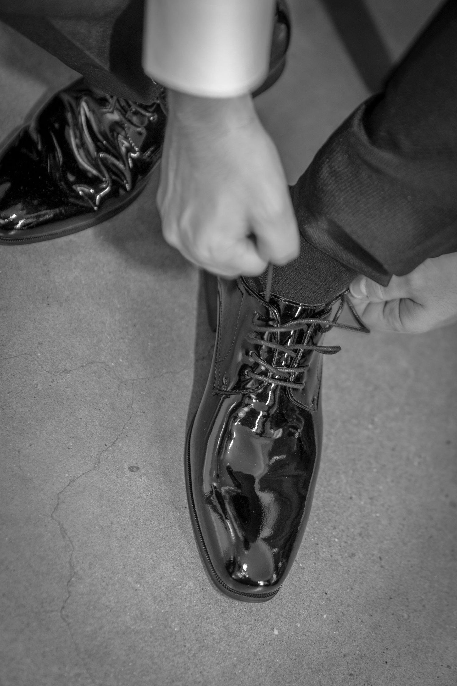 Black and white photo of a man tying shiny patent leather dress shoes in a formal suit.