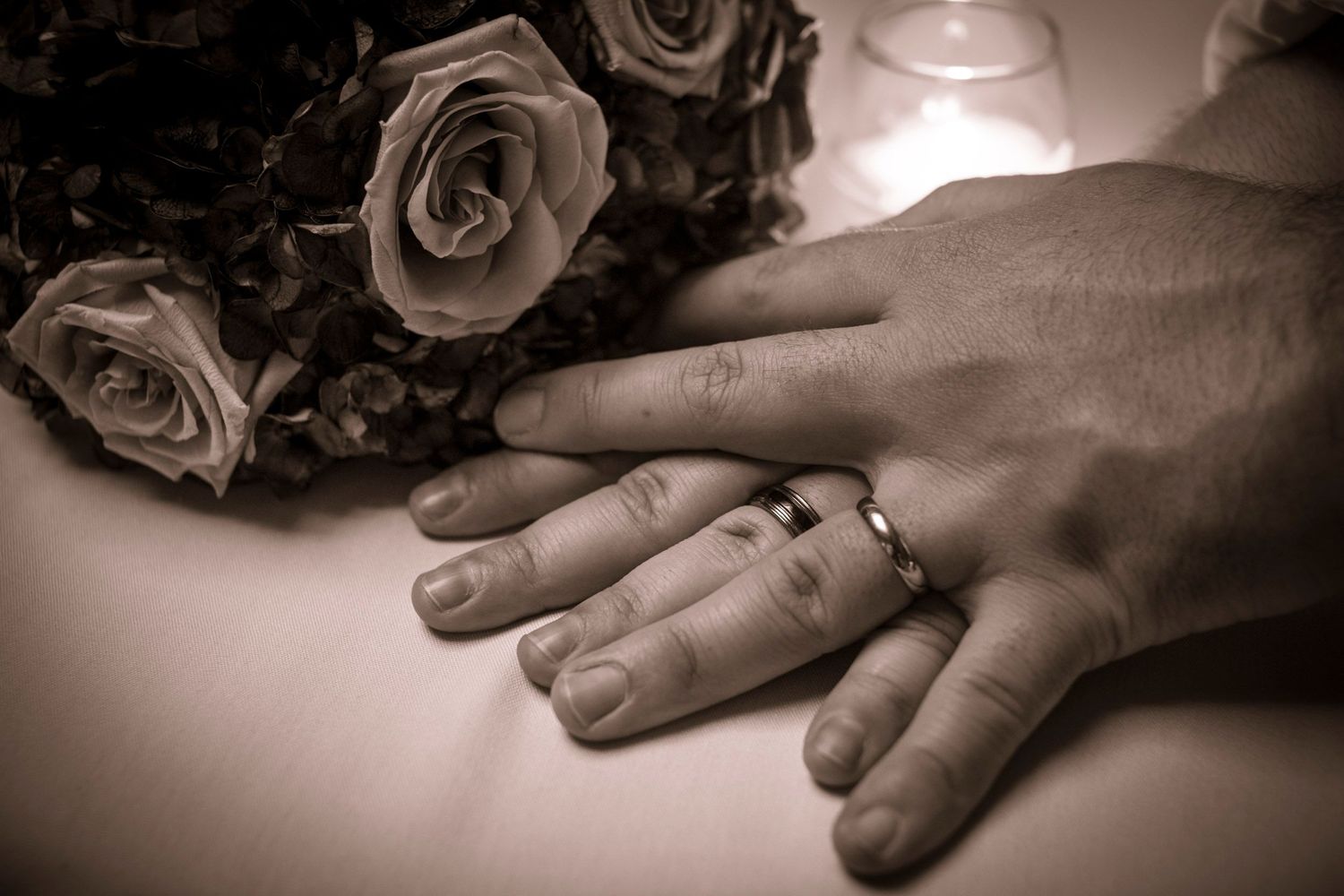 Newlyweds hands with wedding rings resting near roses bouquet in sepia tone.