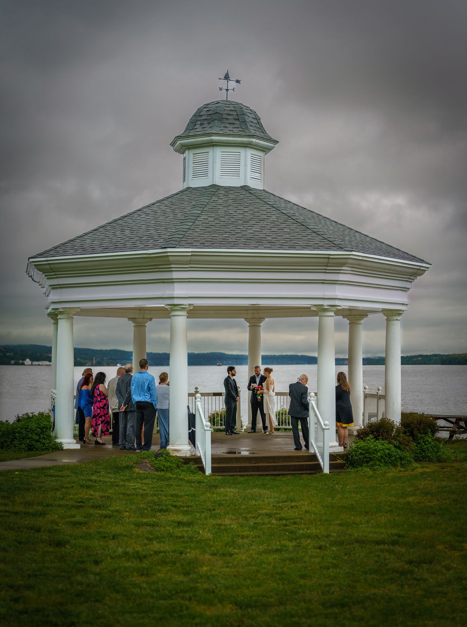Wedding ceremony at a white gazebo overlooking a lake under cloudy skies with guests gathered around.