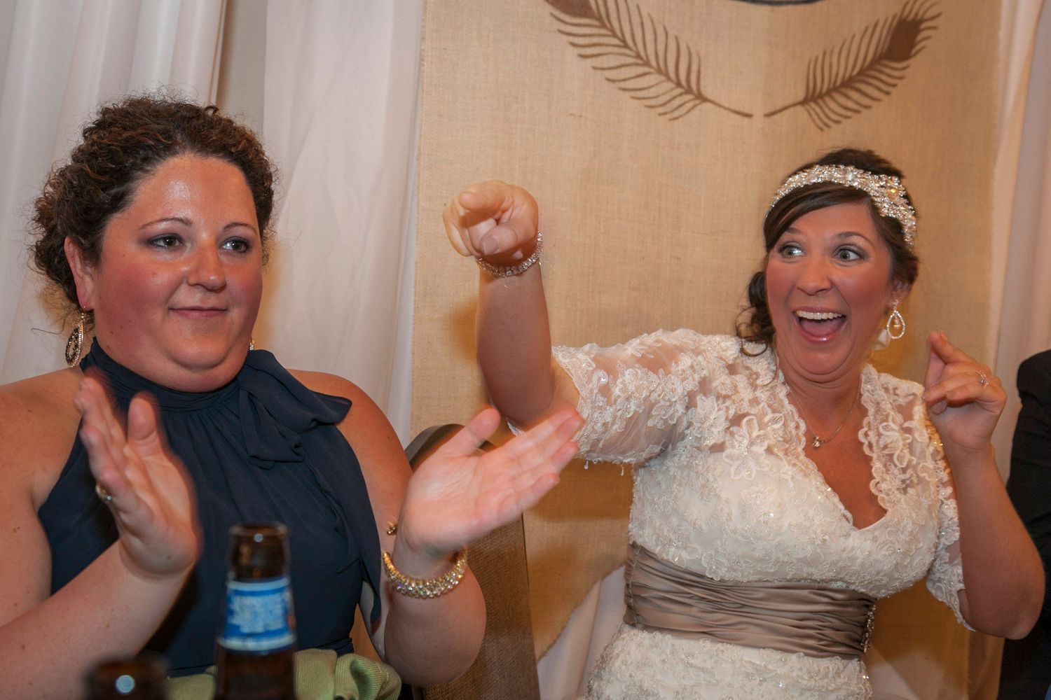 Bride in white lace gown celebrates with bridesmaid in navy dress at wedding reception.