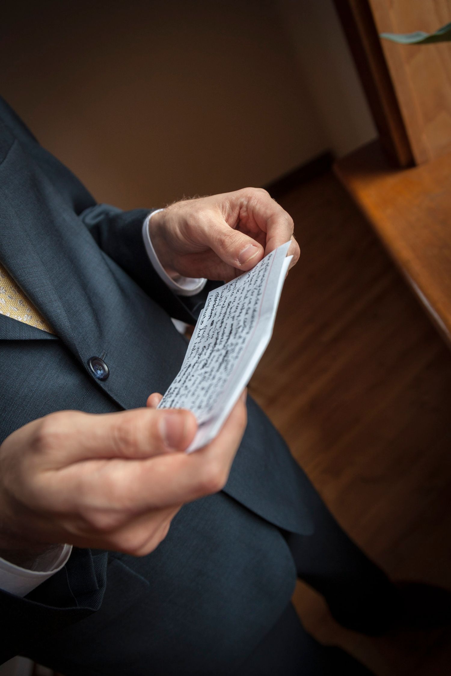 Groom in blue suit reads handwritten wedding vows from folded paper before ceremony.