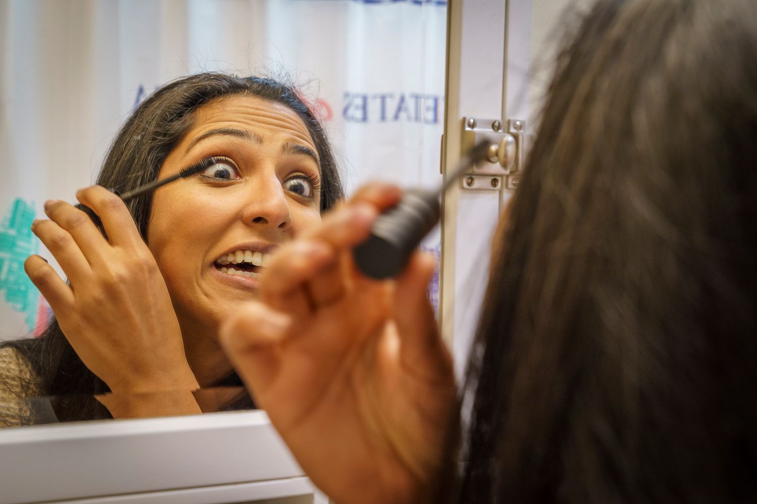 Woman applying makeup with surprised expression in bathroom mirror.