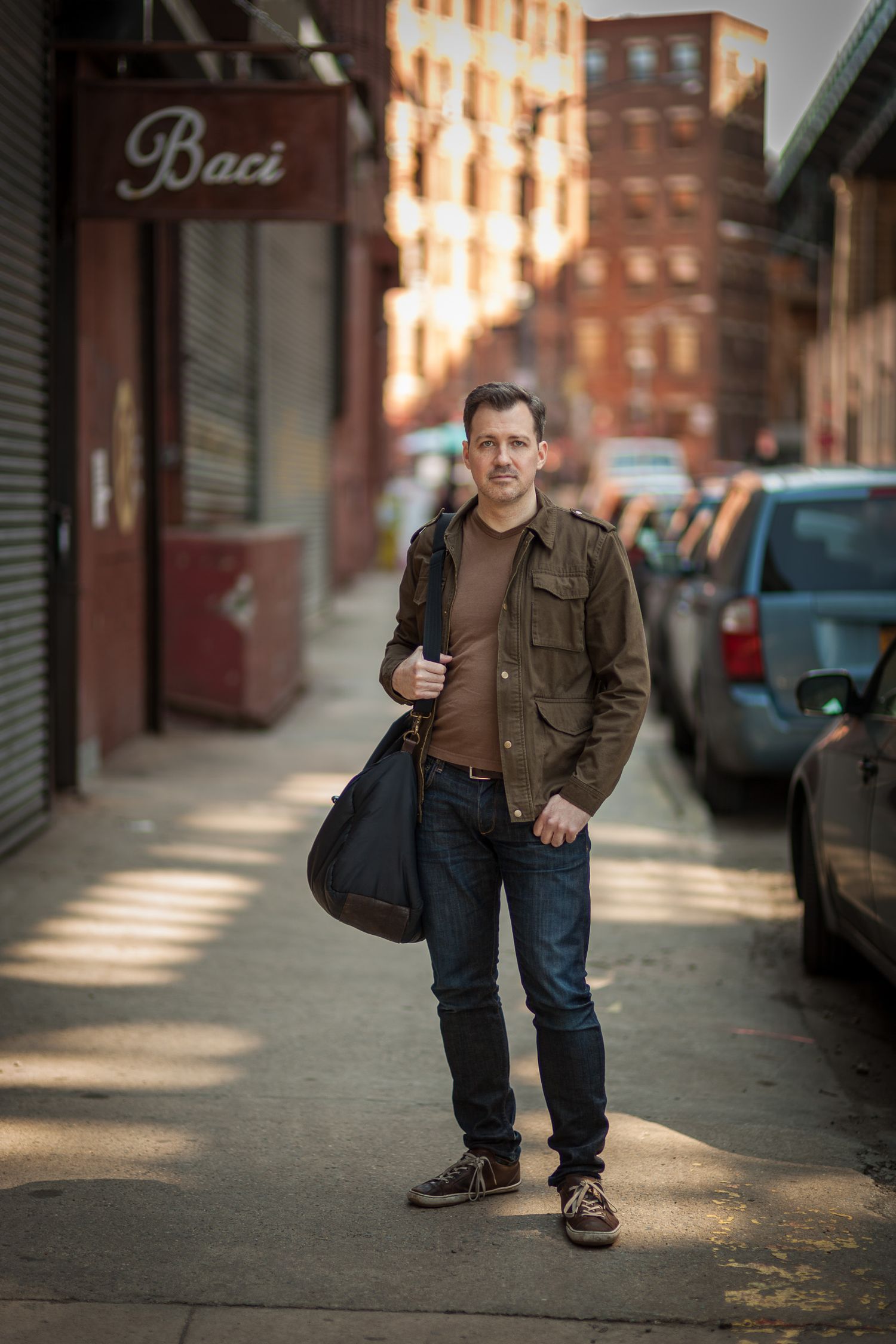 Man with bag standing on urban sidewalk in brown jacket and jeans.