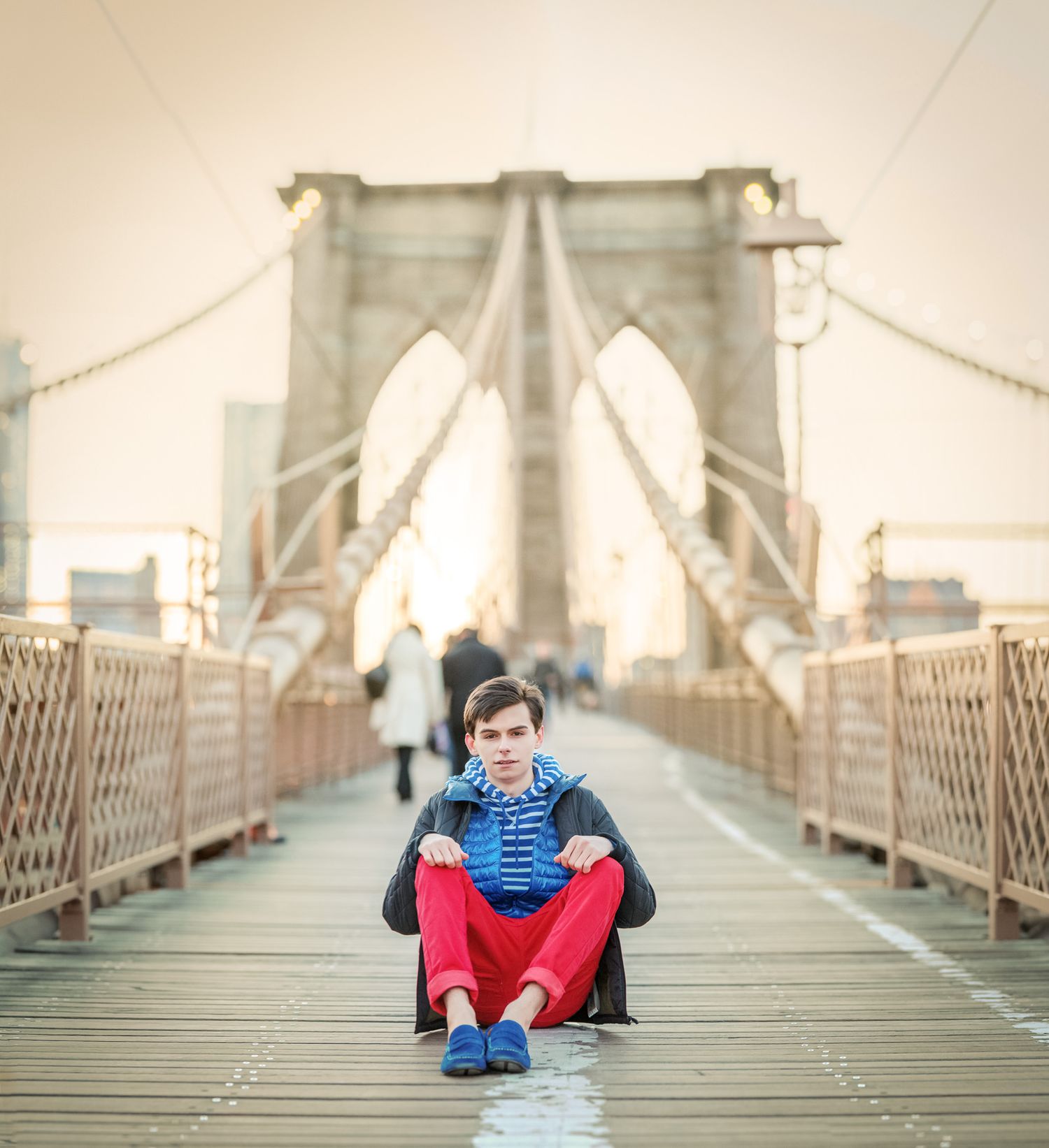 Young man in red pants sitting on Brooklyn Bridge walkway at sunset.