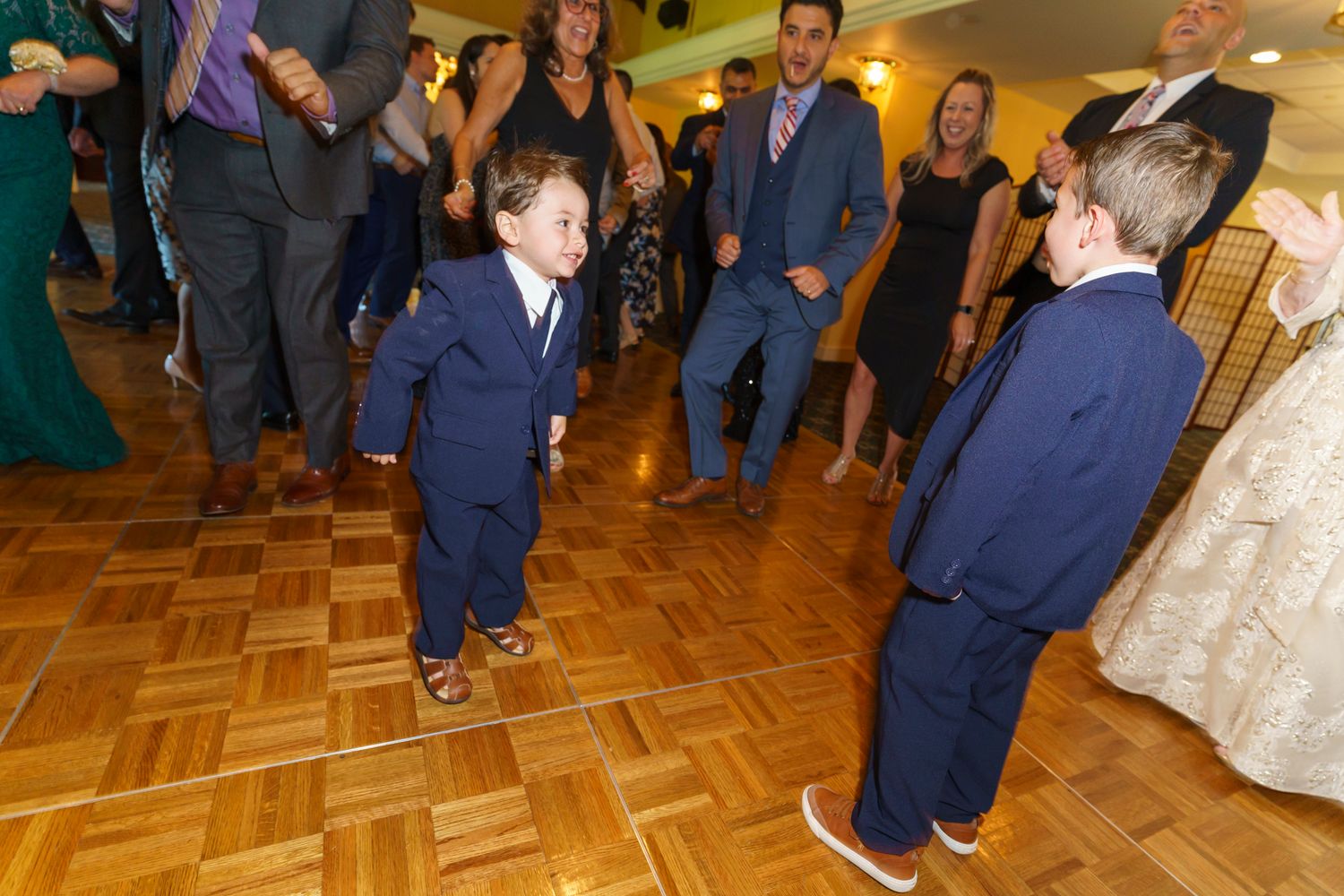 Two young boys in navy suits dance together on a wooden dance floor at a wedding reception.