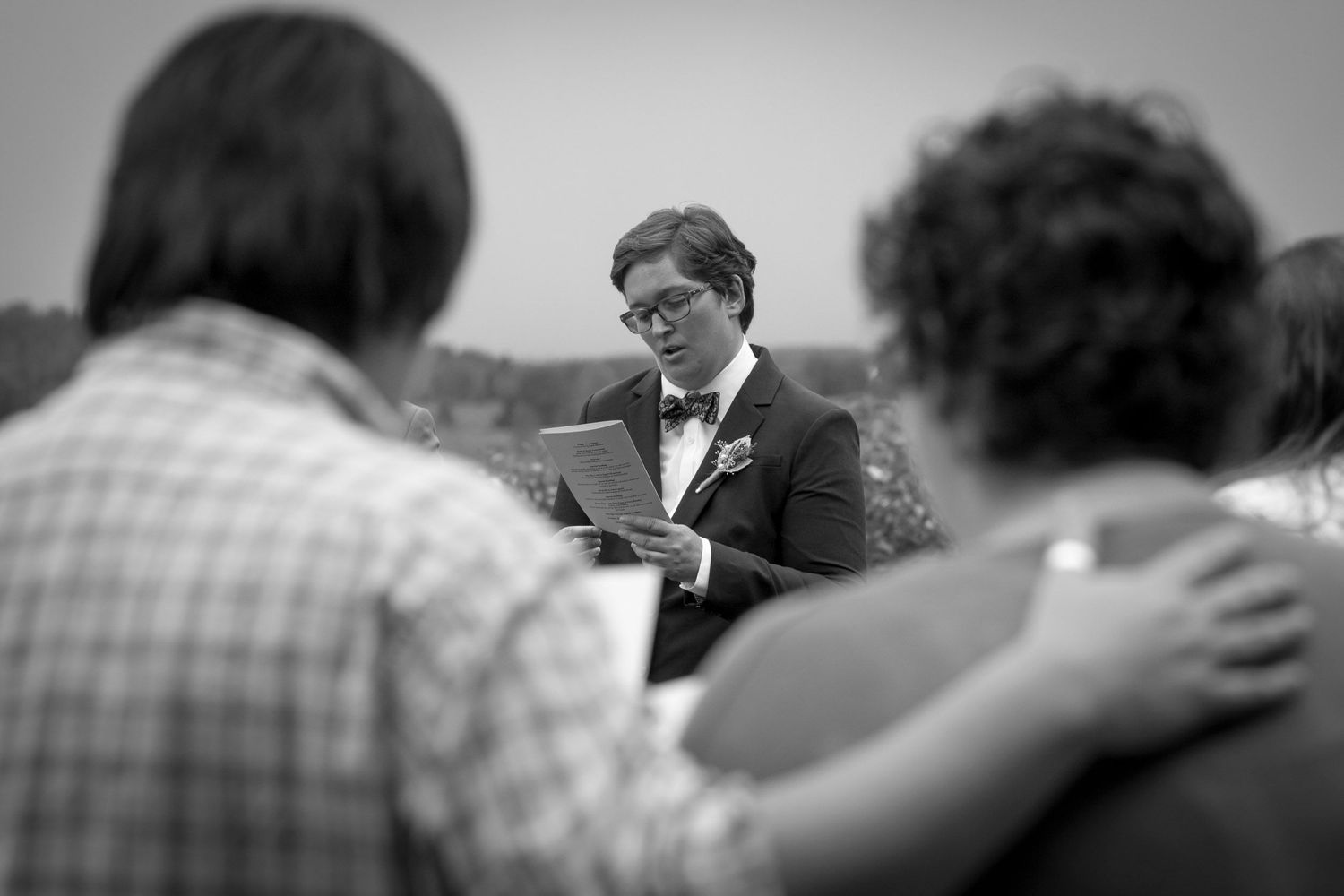 Black and white photo of a man in a tuxedo reading vows at an outdoor wedding ceremony.