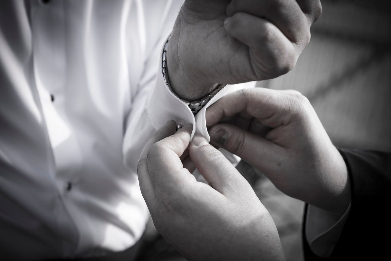 Black and white photo of hands fastening a cufflink on a white dress shirt sleeve.