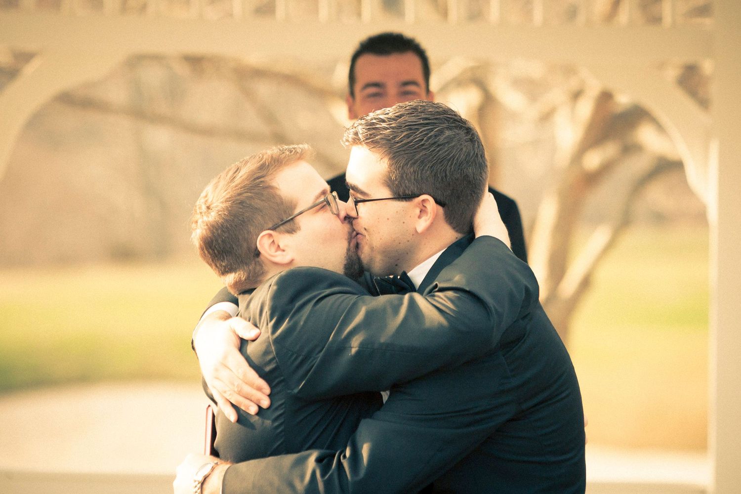 Two grooms sharing a first kiss at their wedding ceremony outdoors near a white gazebo.